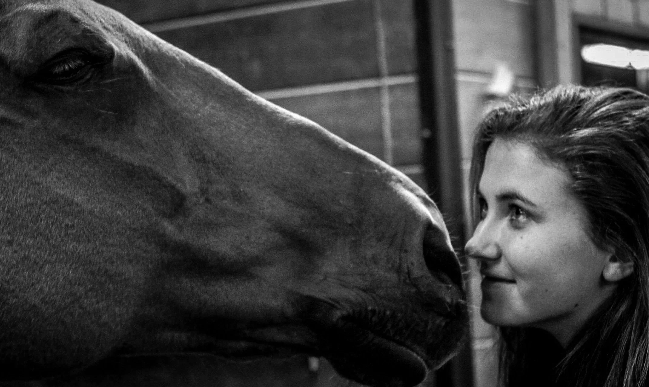 A young woman and a horse face to face indoors, touching noses, in black and white.