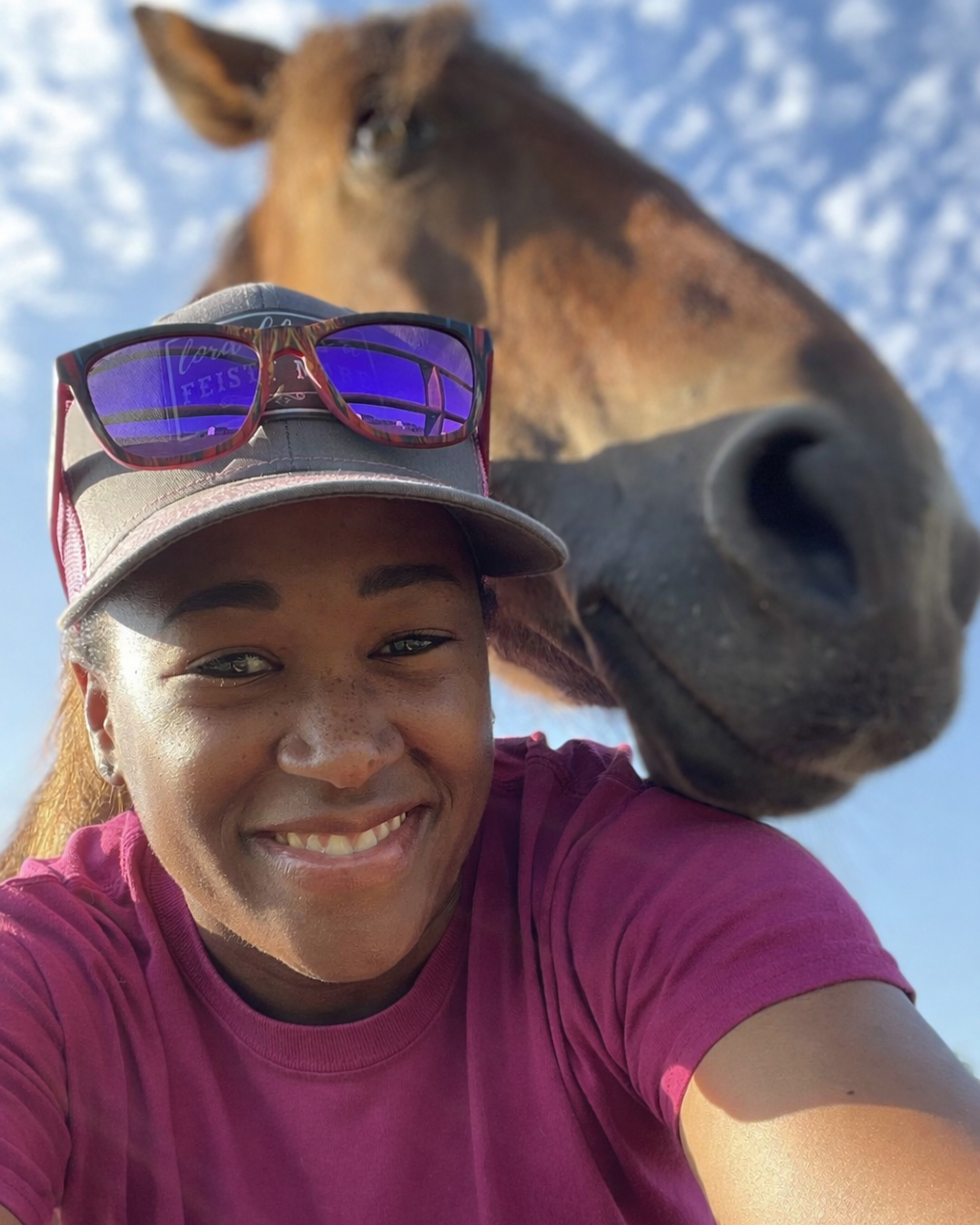 A woman smiling wearing a hat and sunglasses on her head and a purple shirt, with a horse standing behind her