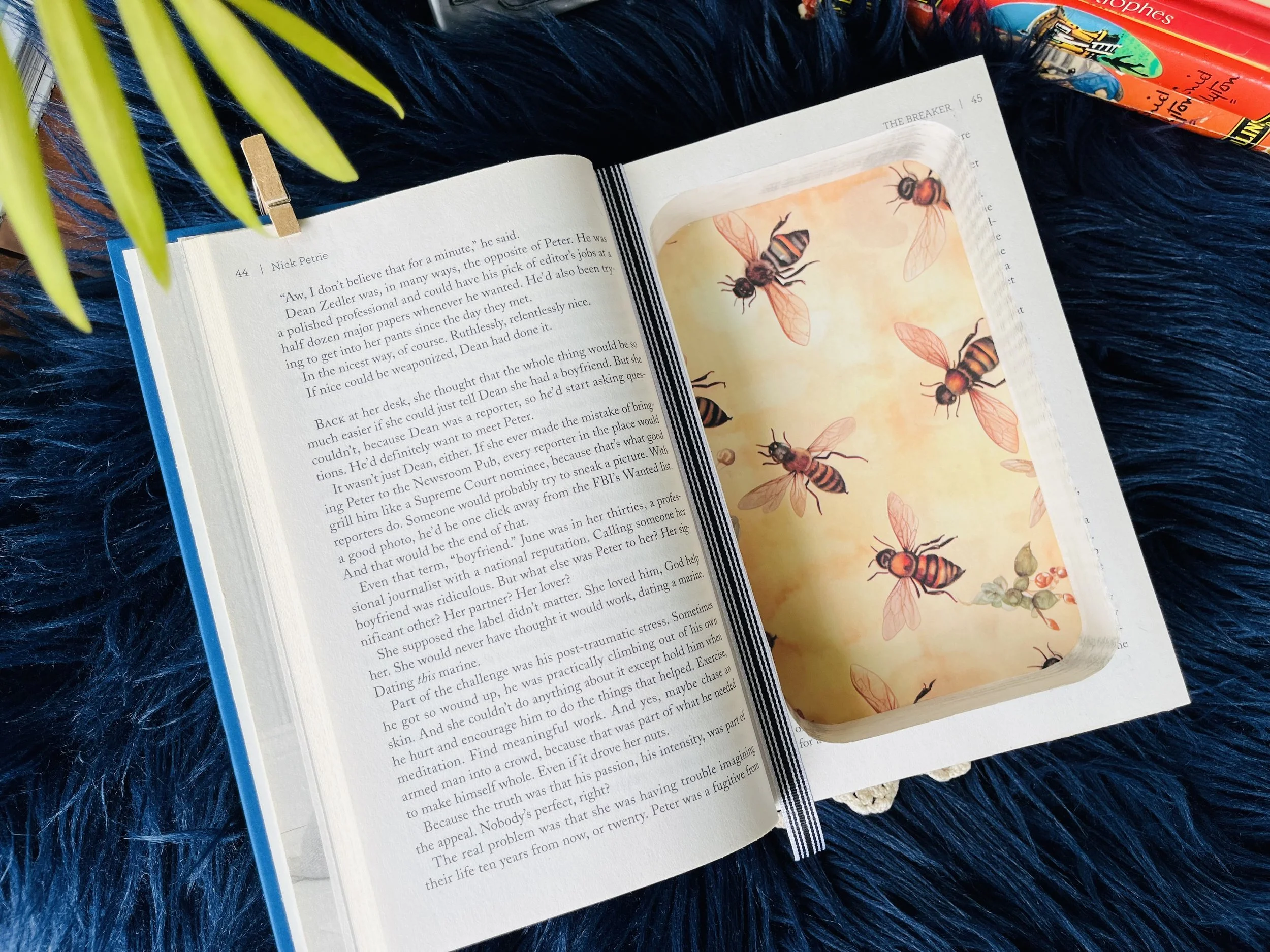 Open book with text on the left page and illustration of bees on the right page, placed on a dark blue textured surface. A green plant leaf is at the top left corner and there are colorful markers at the top right corner.