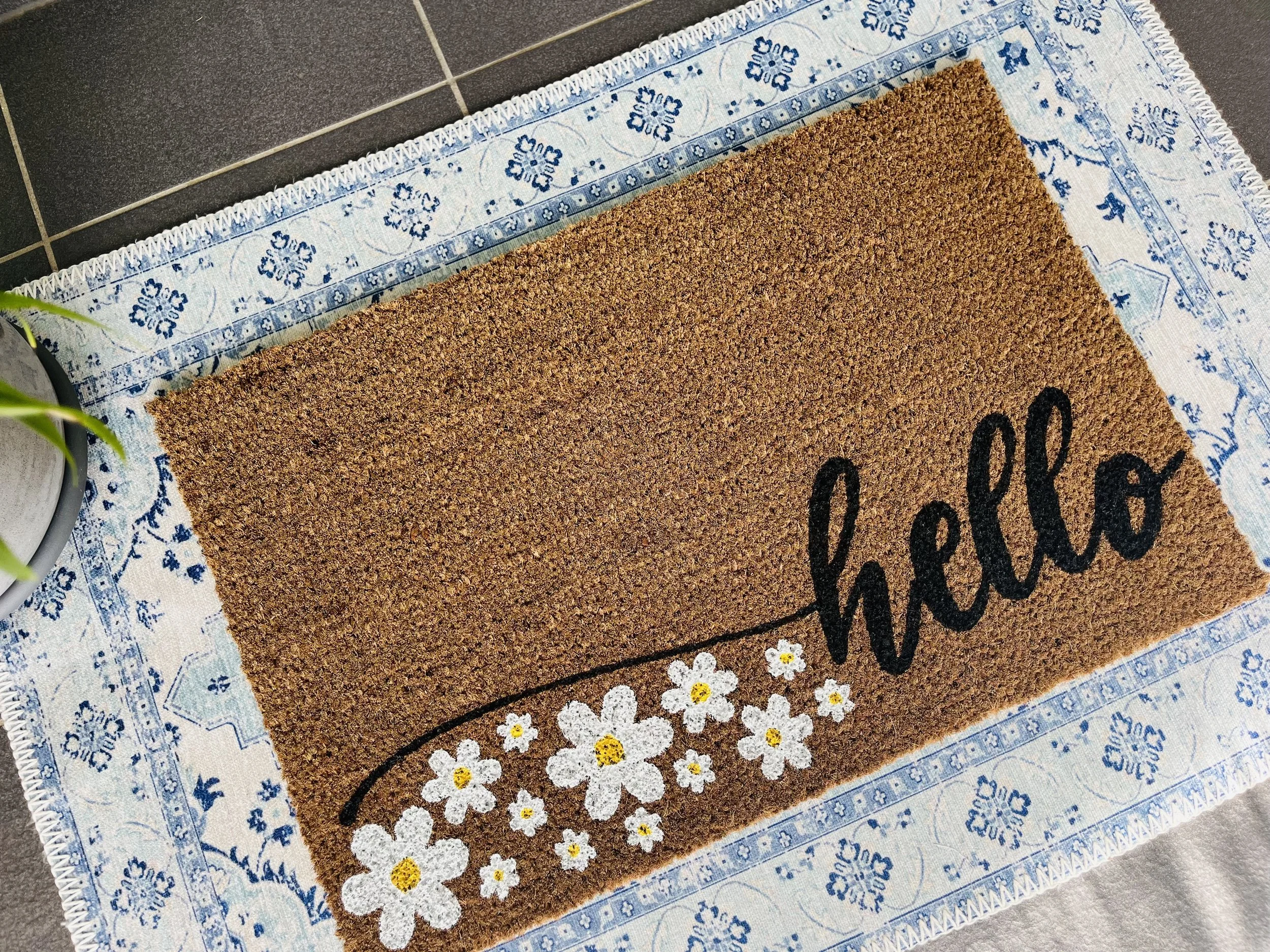 A coir doormat with the word hello written in cursive writing. It has a sweeping H and under that are handpainted white daisies.