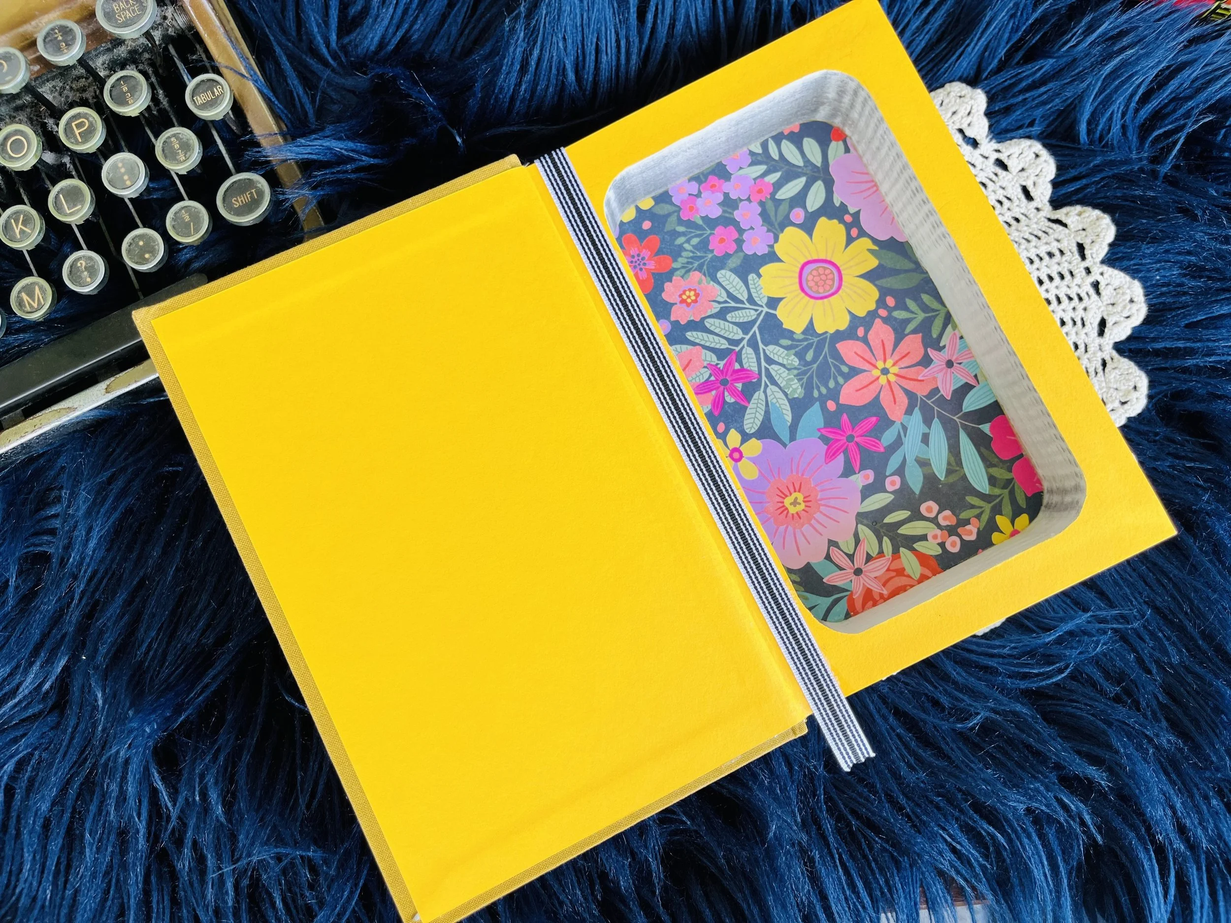 Open yellow box with floral pattern inside, resting on a blue furry surface, next to a vintage typewriter and a white lace doily.