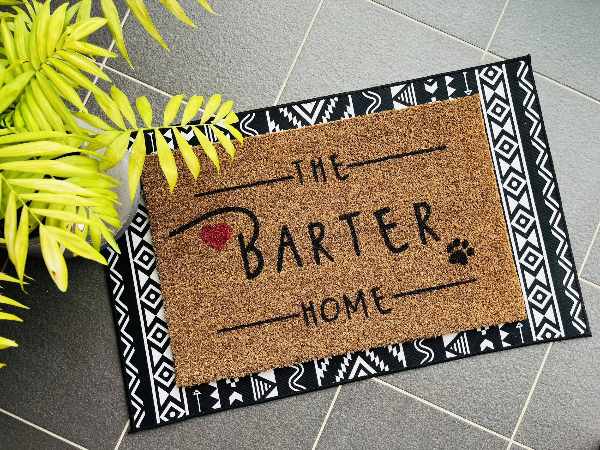 A doormat with the words "The Barter Home" and a red heart, placed on a black and white patterned rug next to a yellow-green plant on a tiled floor.