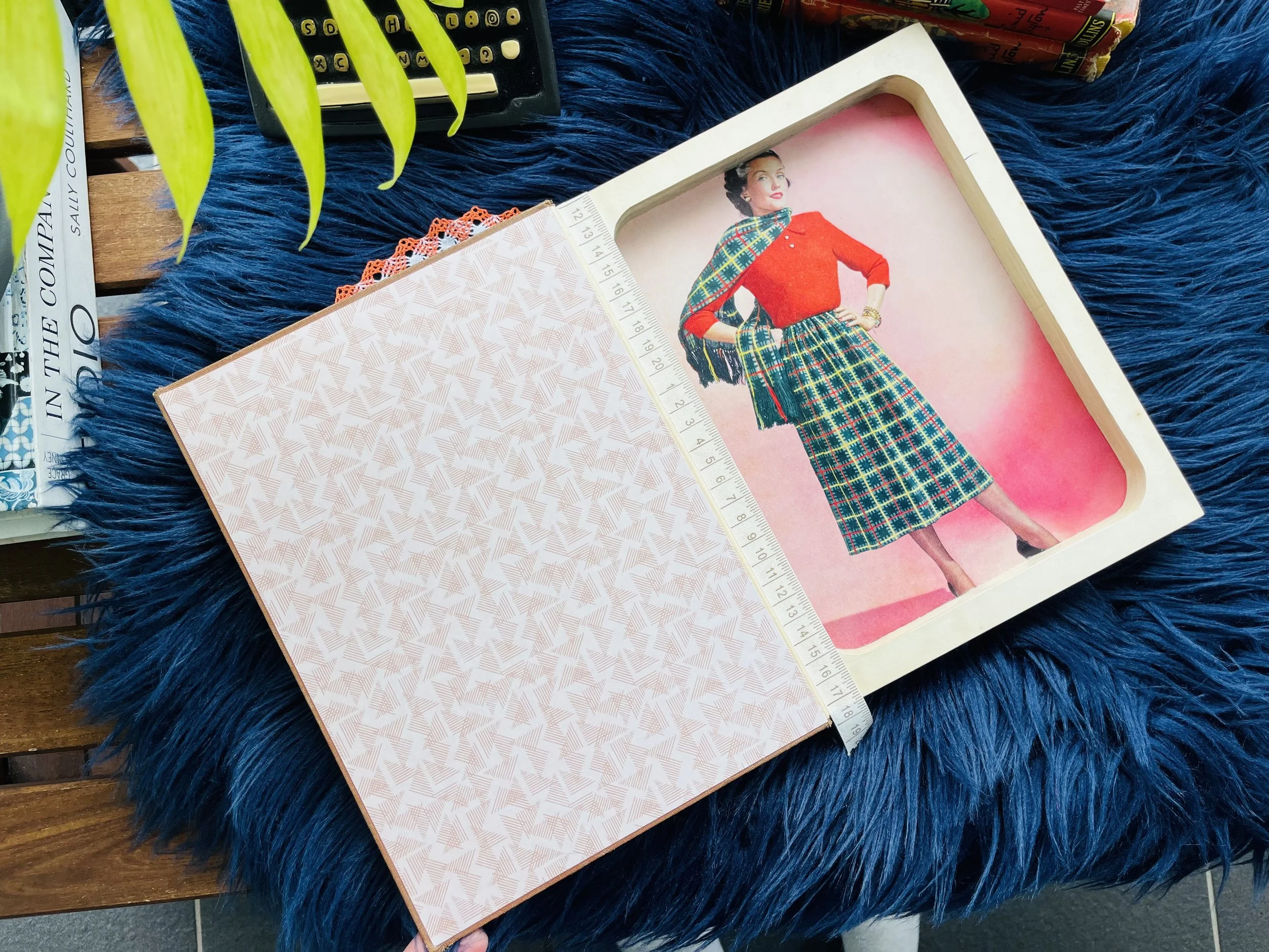 A vintage paper dress pattern with a measuring tape, placed on a fluffy blue surface. The pattern shows a woman in a red blouse and a plaid skirt with a matching plaid scarf. Part of a plant, a magazine, and some books are visible nearby.