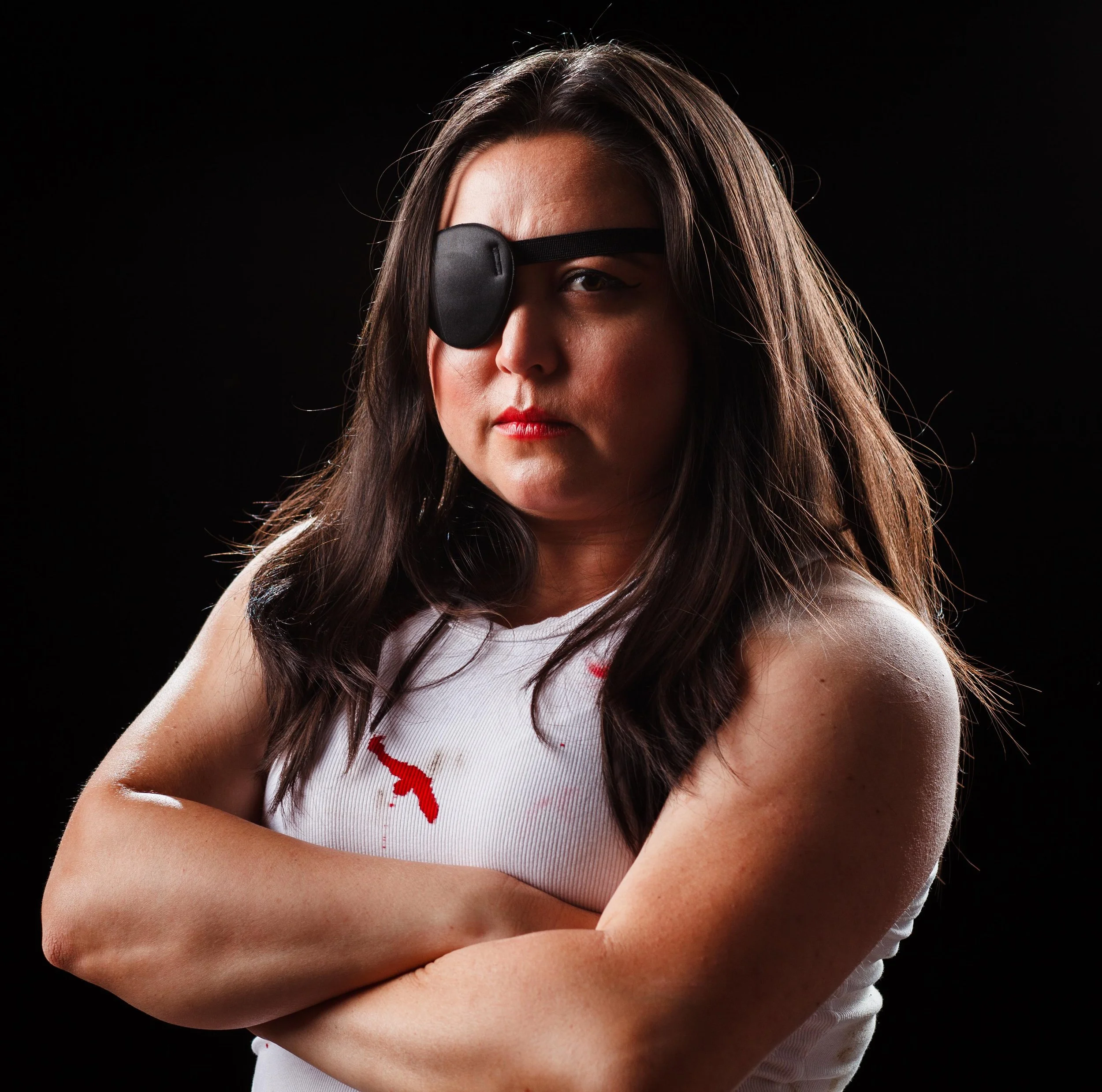 A woman with long dark hair wearing a black eye patch over her right eye, standing with arms crossed against a black background.