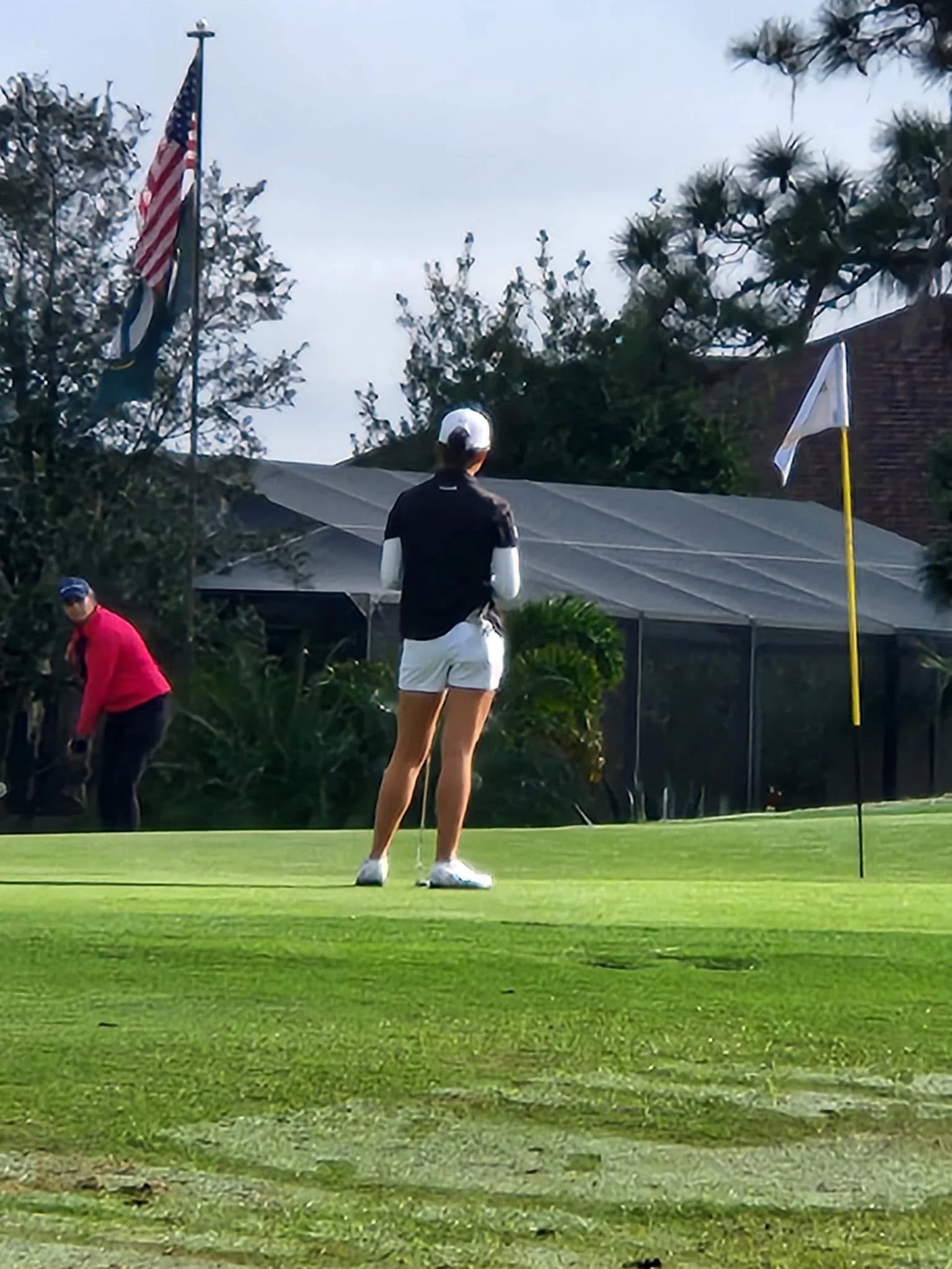 A golfer stands near the hole on a green, with a flag on a yellow pole. Another person in a pink top is in the background, near trees and a fence. An American flag is flying on a flagpole.