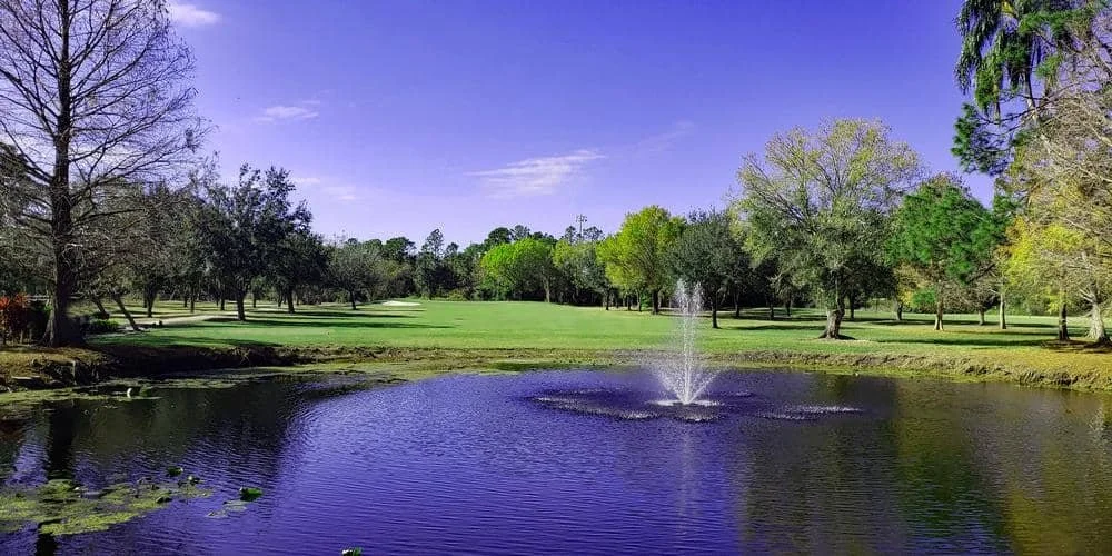 A scenic view of a golf course with a pond and fountain in the center, surrounded by trees and a blue sky.