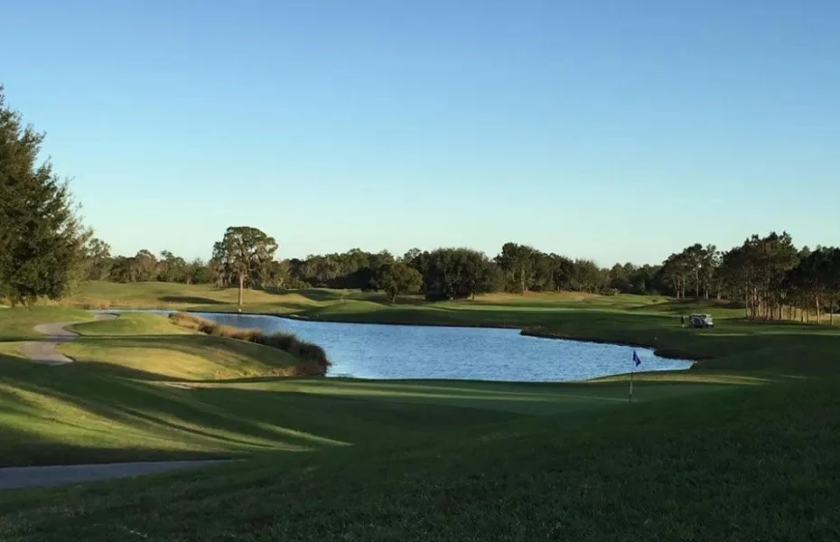 A golf course with a water hazard, green grass, trees, and a golf flag.
