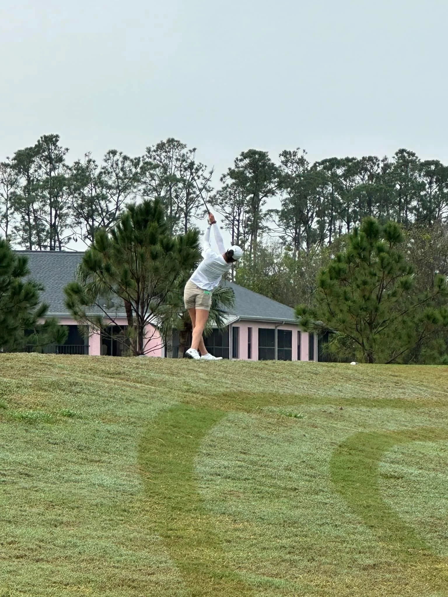 A person in a white shirt and beige shorts swinging a golf club on a golf course.