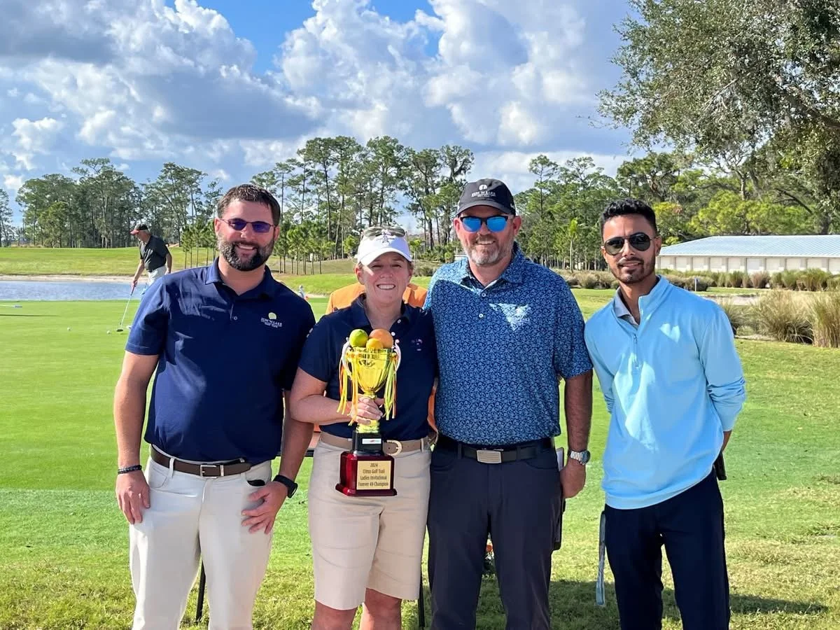 Group of four people standing on a golf course, smiling, with a woman holding a trophy with golf balls on top. Background shows trees, a pond, and a person putting.