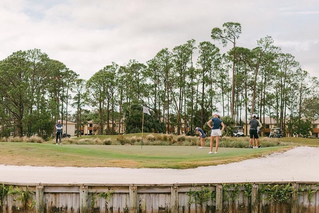 Four people playing golf on a course surrounded by trees and residential buildings, with a sand trap and a flag on the green in the foreground.