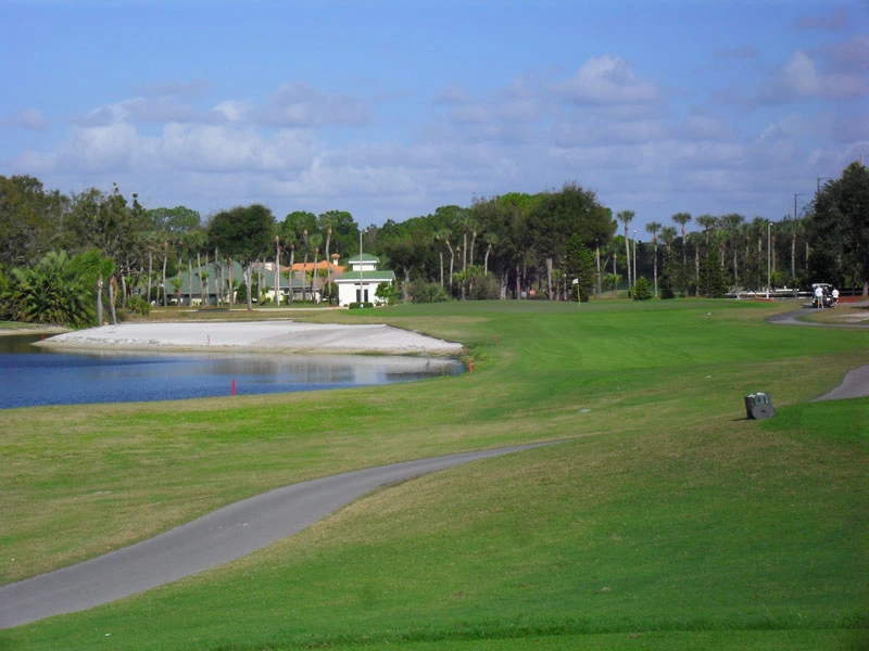 A golf course with a small water hazard, a sand trap, green grass, trees, and a building with a white exterior in the background under a partly cloudy sky.