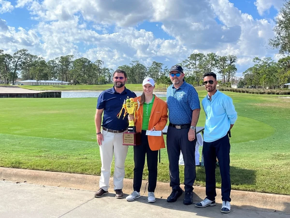 Group of four people standing on a golf course, holding a trophy and check, with trees and a pond in the background on a sunny day.