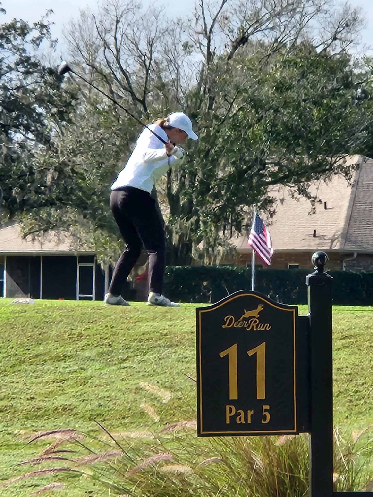 A woman in a white shirt and black pants playing golf on a grassy course, preparing to hit the ball near a sign that reads "Deer Run, hole 11, Par 5."