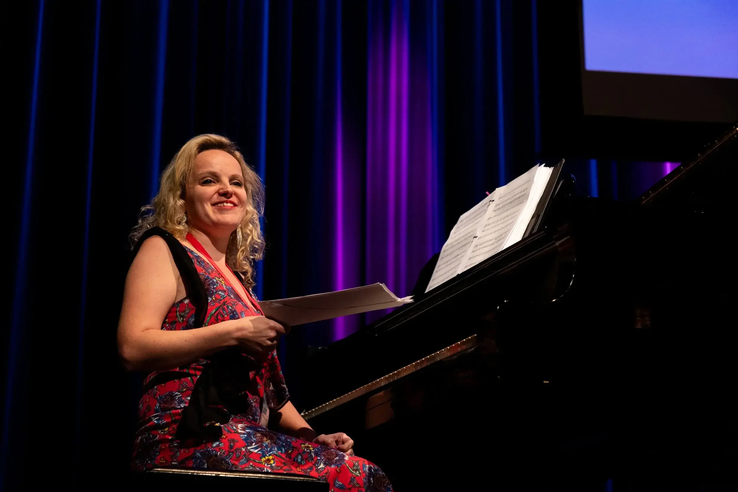 A young woman with long curly hair and glasses, dressed in a black suit, smiling and holding two flutes, standing in front of a dark background.