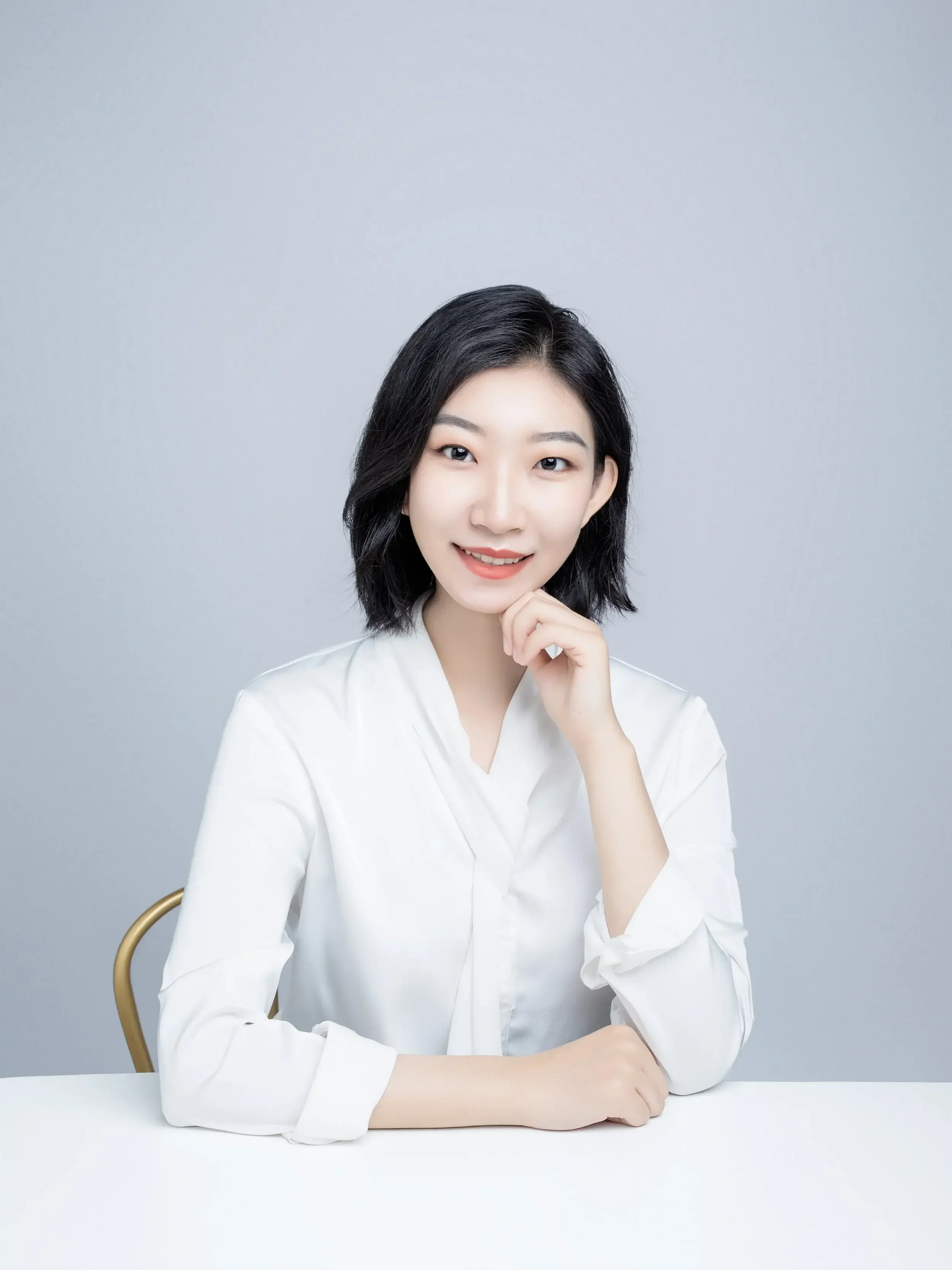 A young woman with black, shoulder-length hair, smiling and resting her chin on her right hand, seated at a white table against a light gray background
