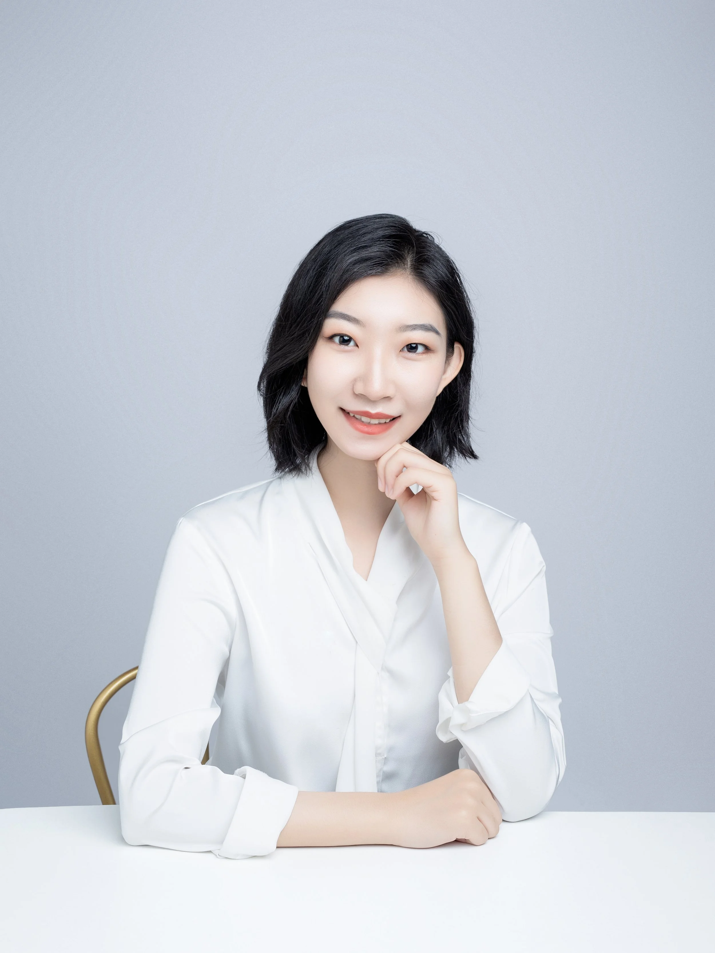 A young woman with black, shoulder-length hair, smiling and resting her chin on her right hand, seated at a white table against a light gray background