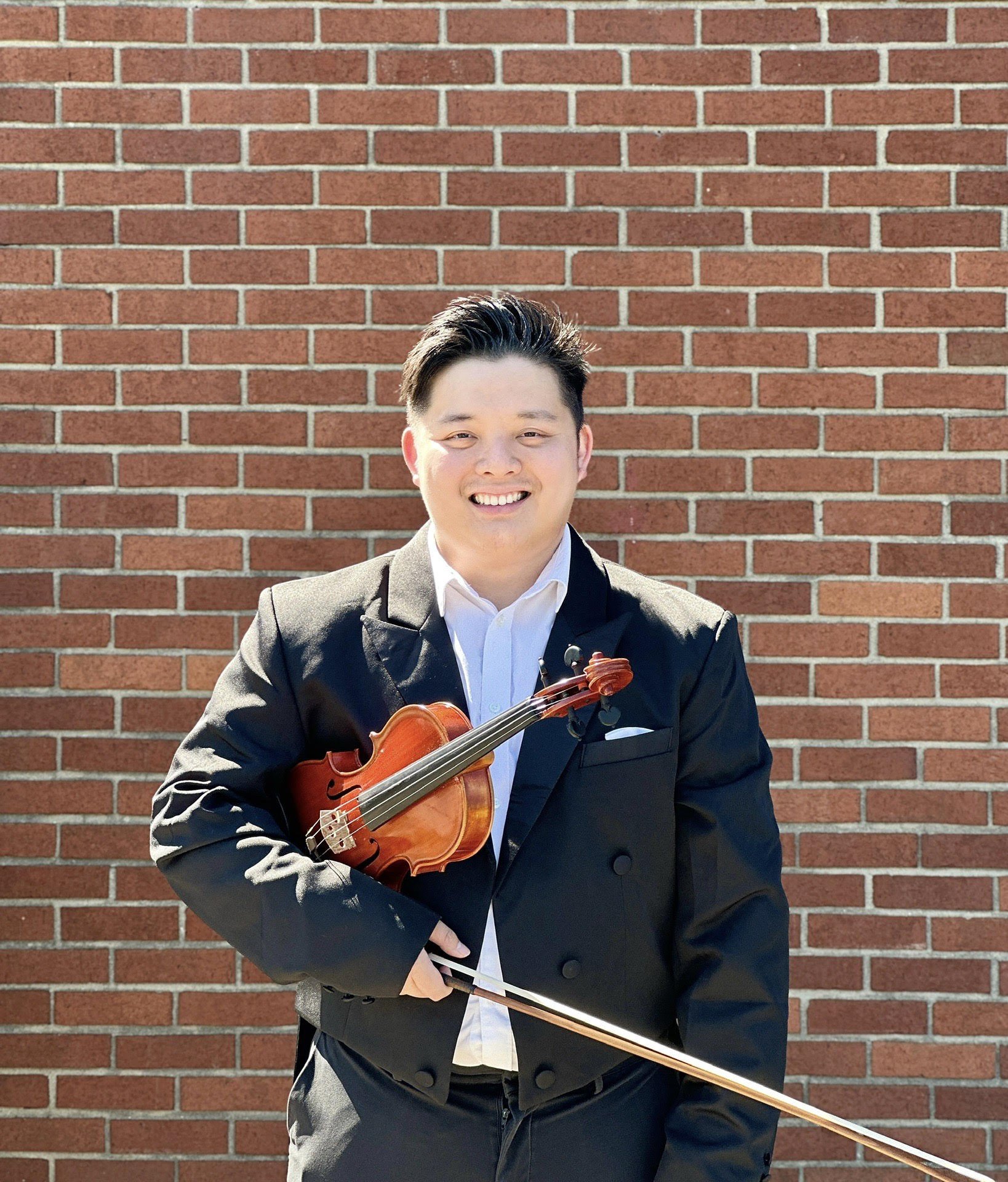 A young man in a black suit holding a small wooden violin and a bow, standing in front of a brick wall, smiling at the camera.