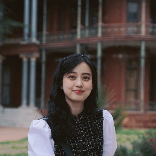 A young woman with long black hair and a white shirt with a black vest stands outdoors in front of a historic building with iron balconies and stone steps.