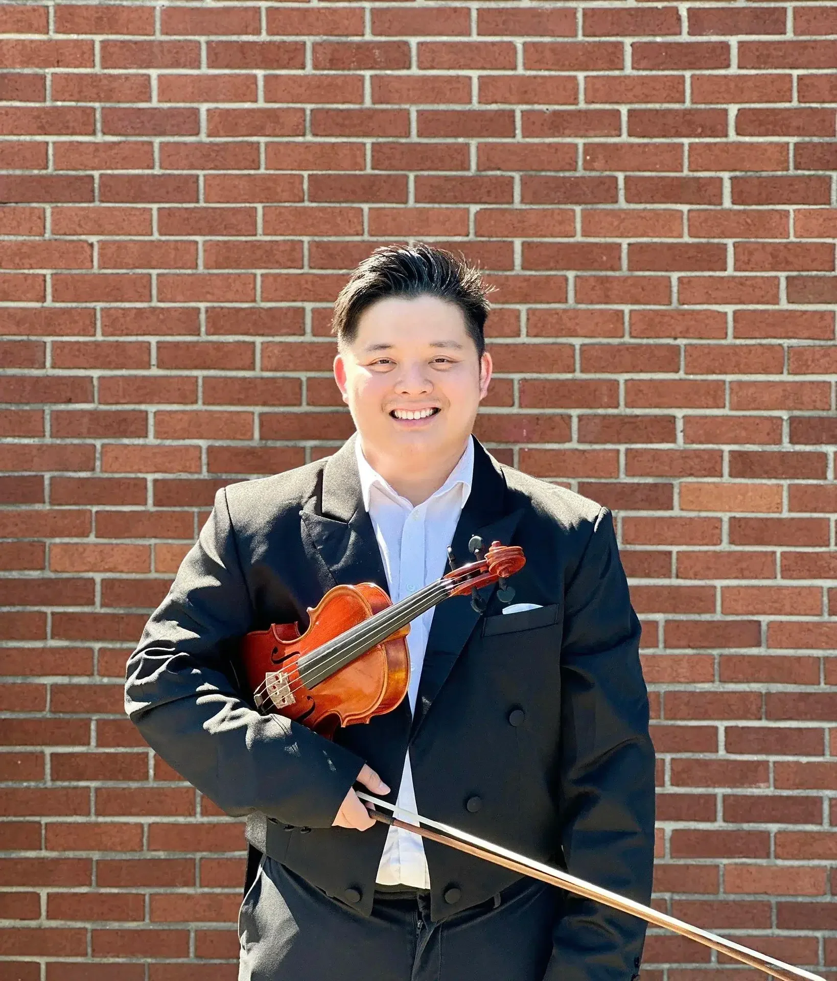 A young man in a black suit holding a small wooden violin and a bow, standing in front of a brick wall, smiling at the camera.