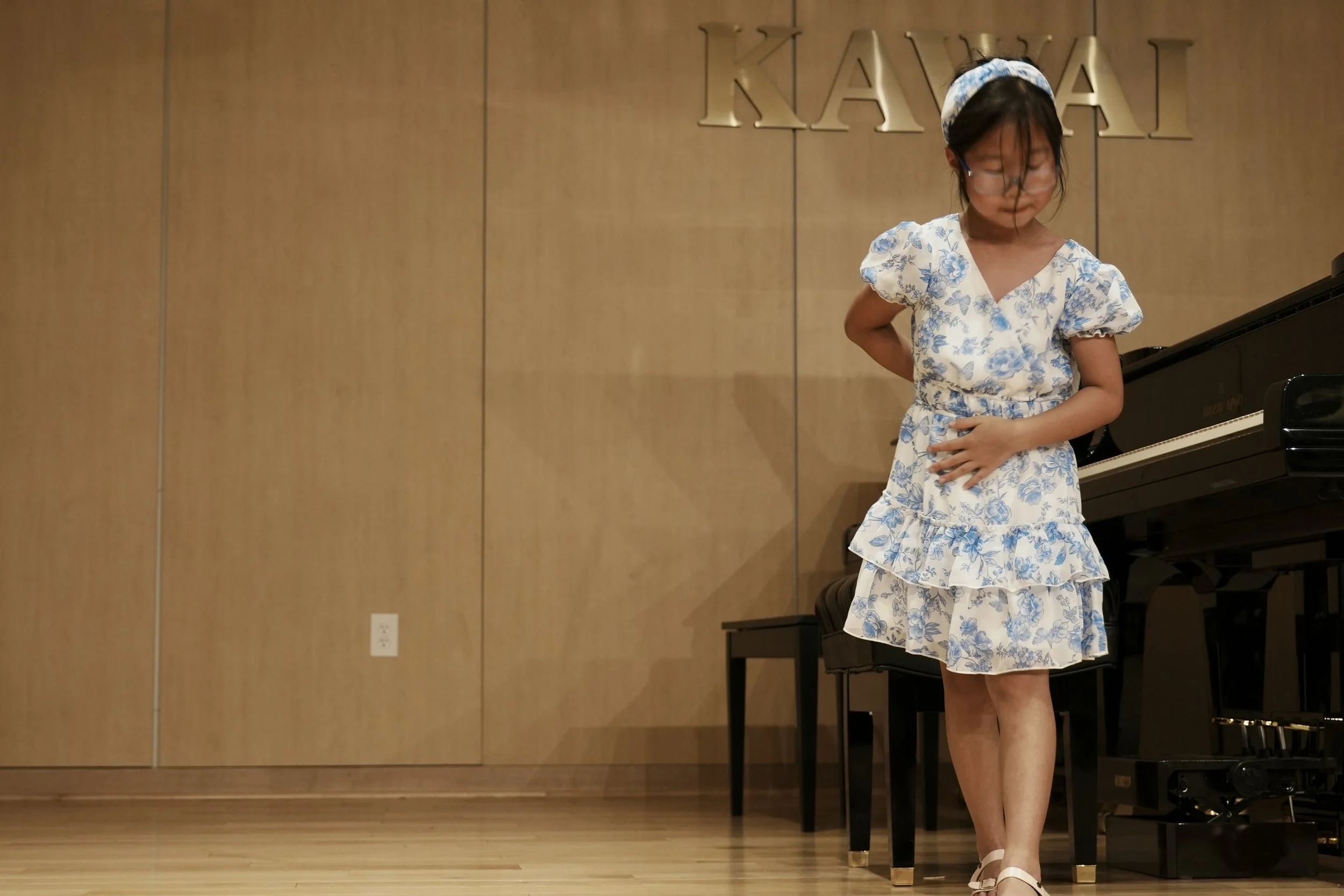 A young girl in a white and blue floral dress standing next to a black piano on a wooden stage, with the word 'KAVAI' in gold letters on the wooden wall behind her.