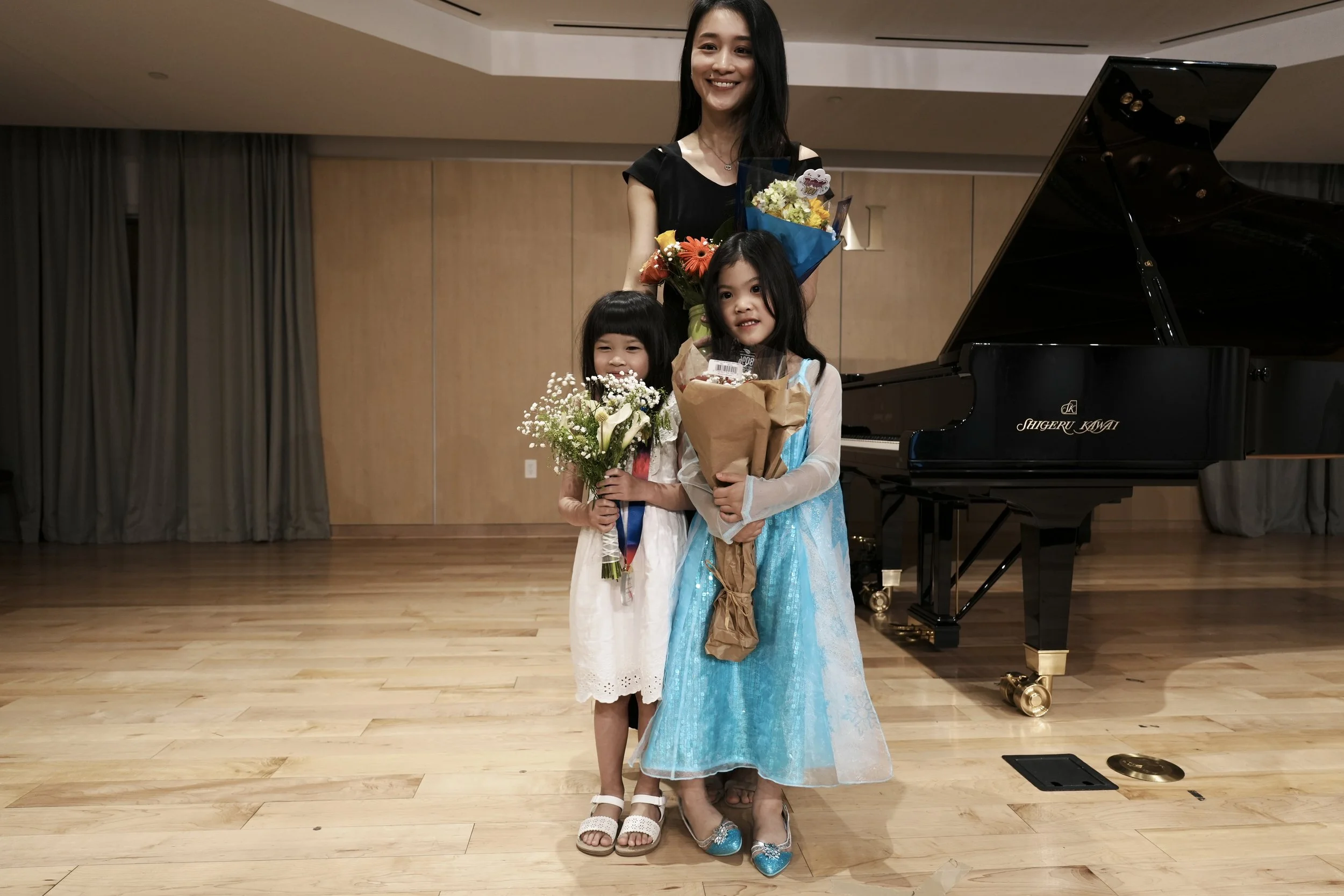 A woman and two young girls standing in front of a black grand piano on a wooden stage. The women and girls are holding flowers and looking at the camera, smiling. The woman is positioned behind the girls, who are in the front, with the girls holding