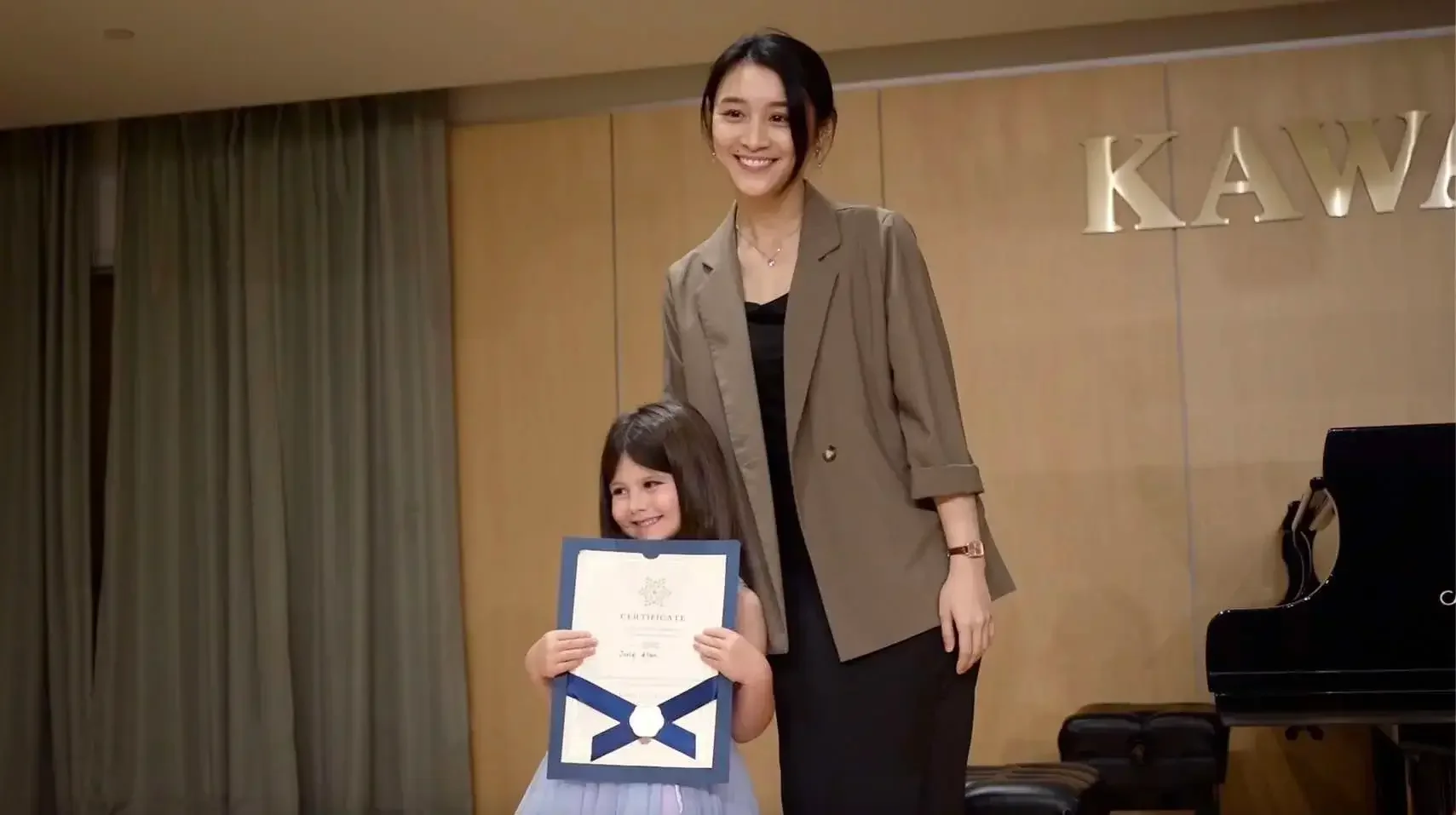 A woman and a young girl standing together, smiling, with the girl holding a certificate, in a room with a piano and beige curtains.