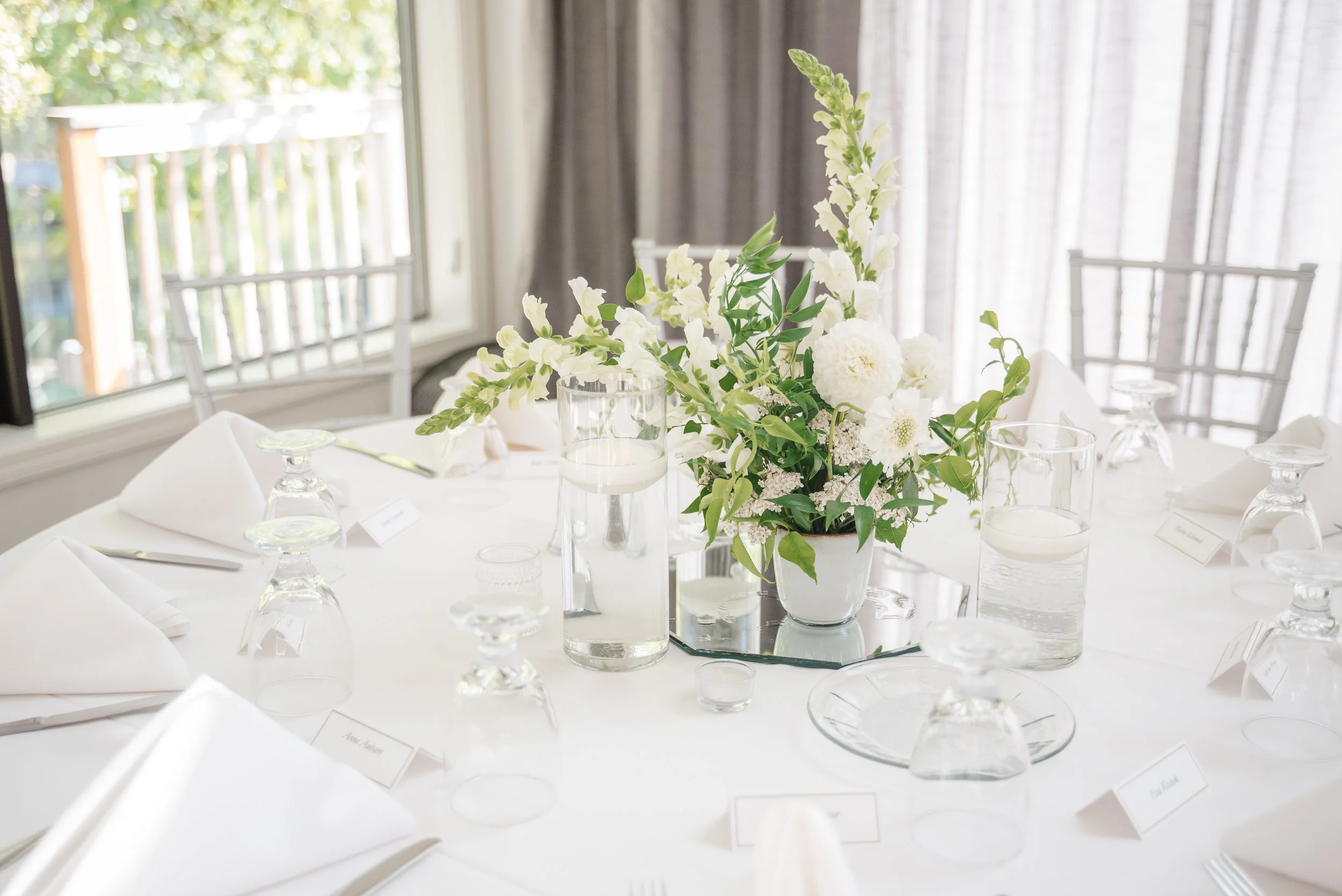 Elegant dining table with a large floral centerpiece in a white vase, surrounded by water glasses, candles, and folded white napkins, in a well-lit room with large windows and curtains.