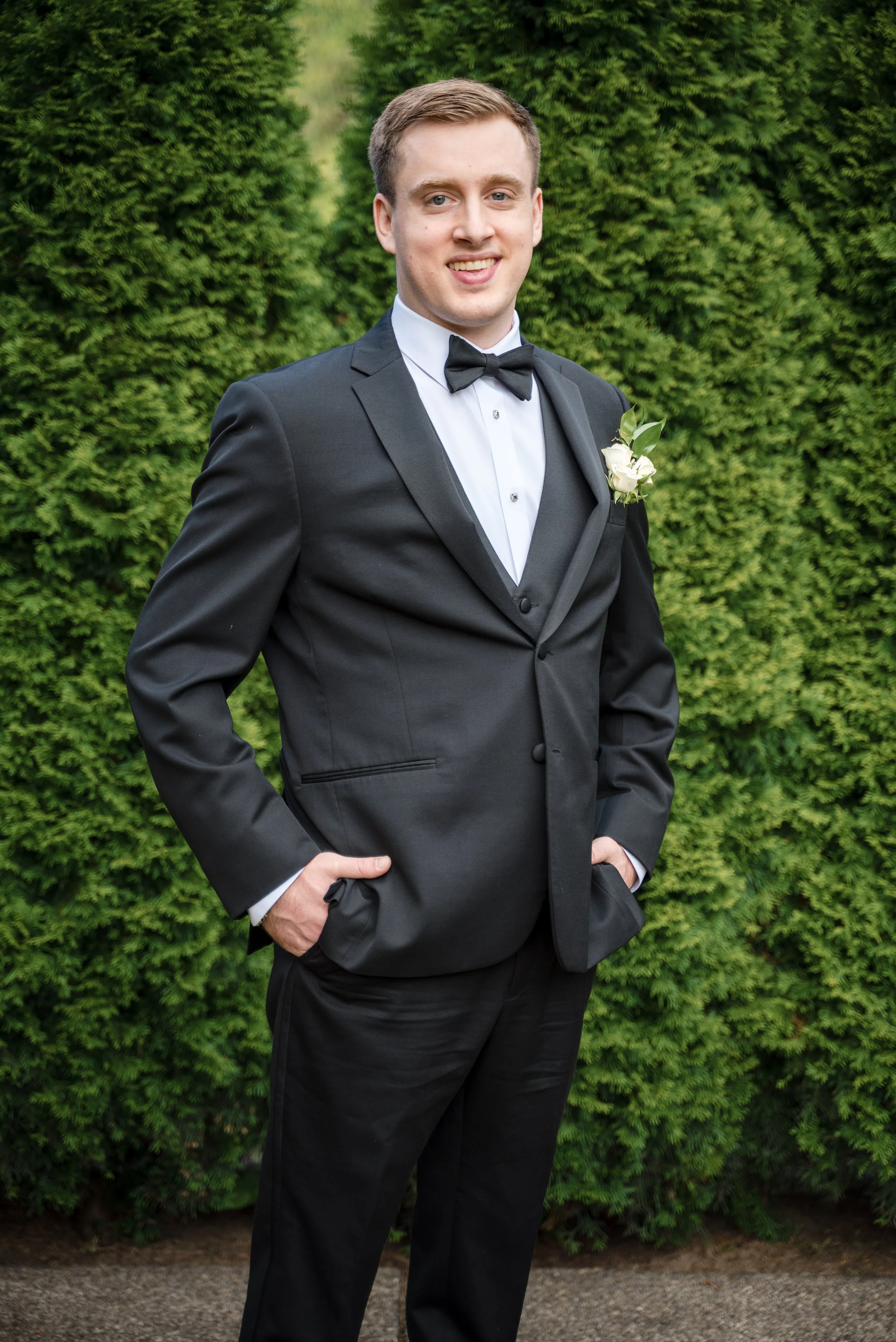 A young man dressed in a formal black tuxedo with a white dress shirt, black bow tie, and boutonniere, standing outdoors in front of green foliage.