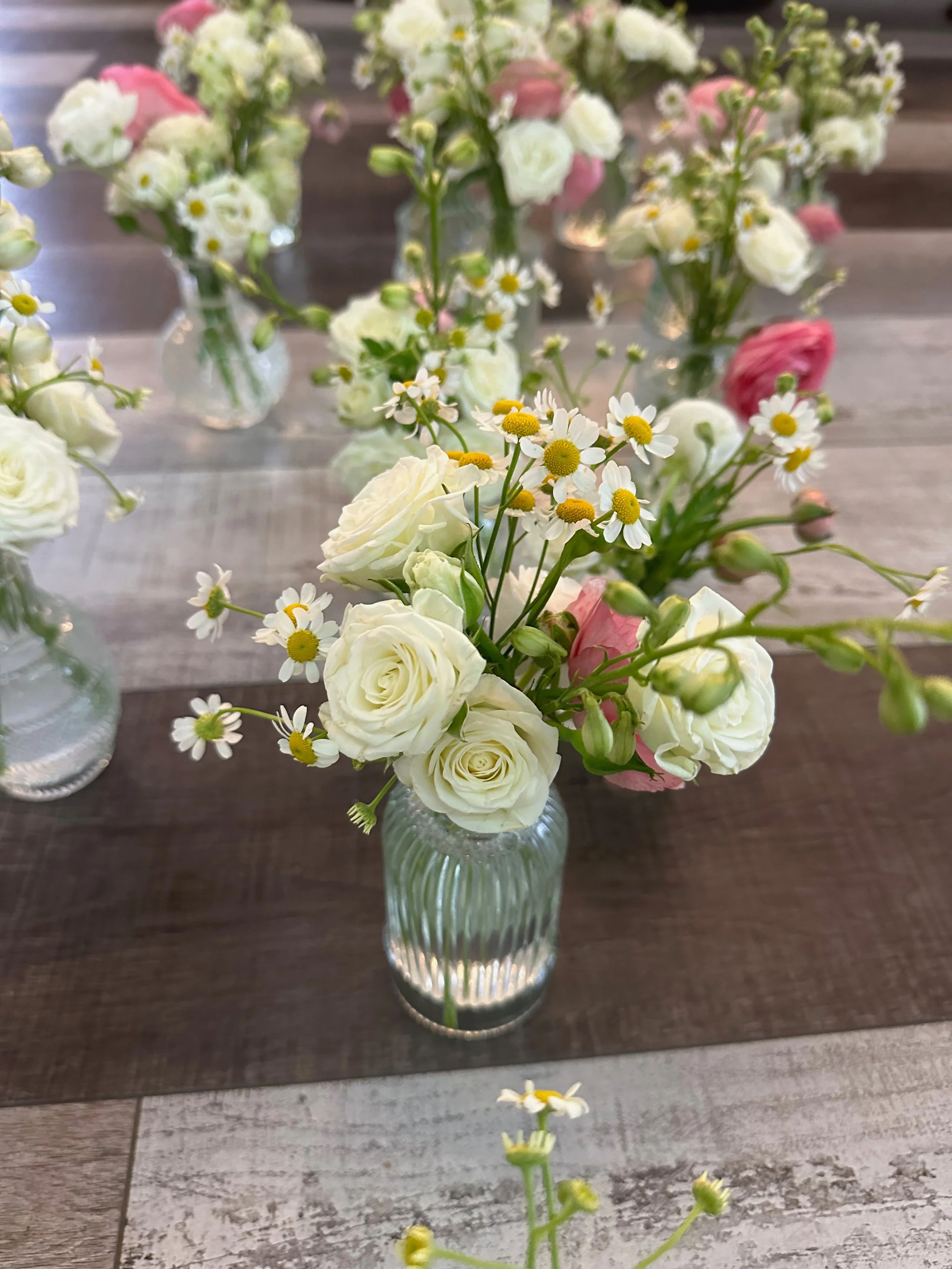 Small glass vase with a floral arrangement of white roses, daisies, and pink flowers placed on a wooden table.