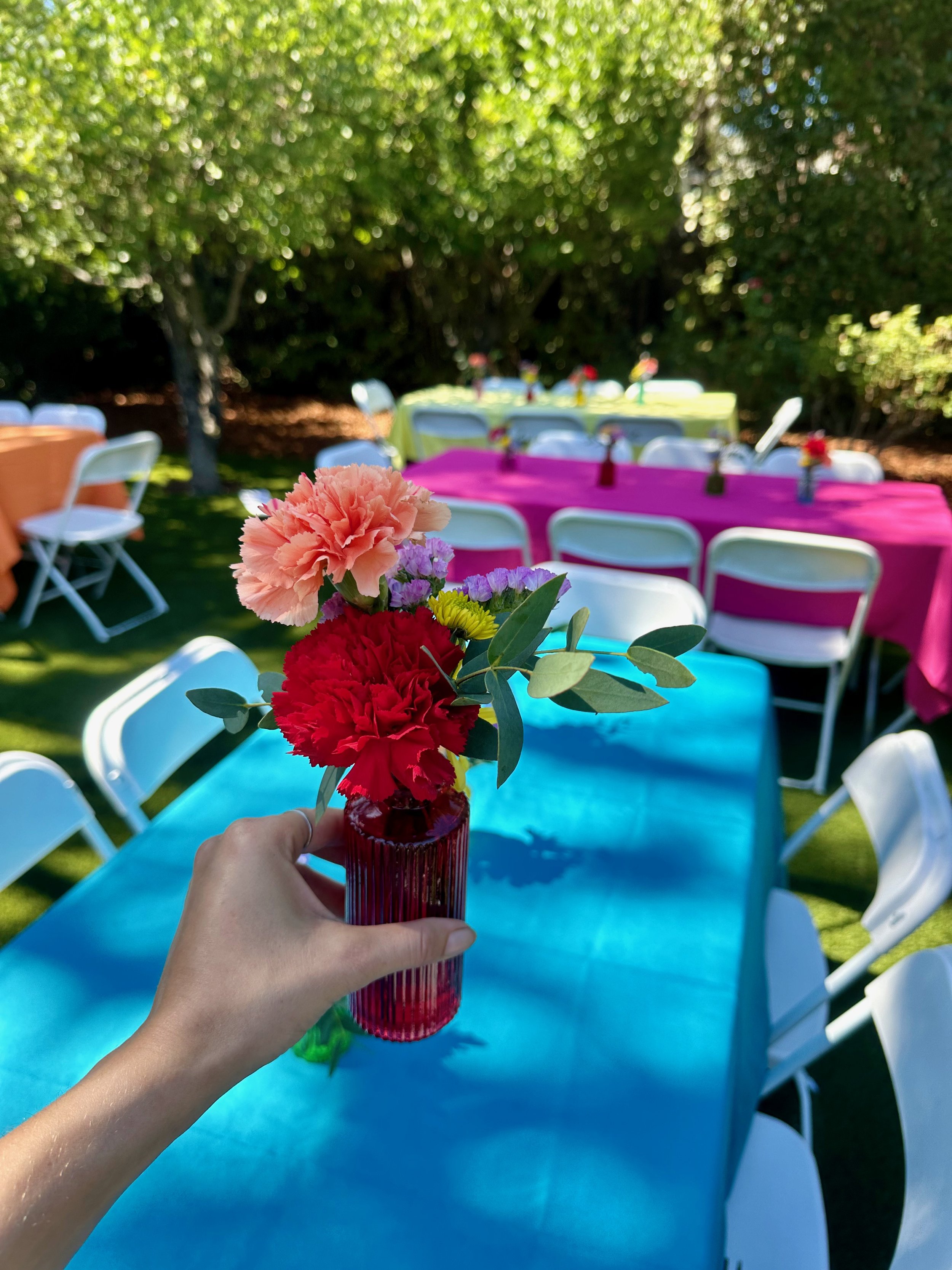 Person holding a red vase with pink, red, purple, and yellow flowers, set on a blue table at outdoor event.