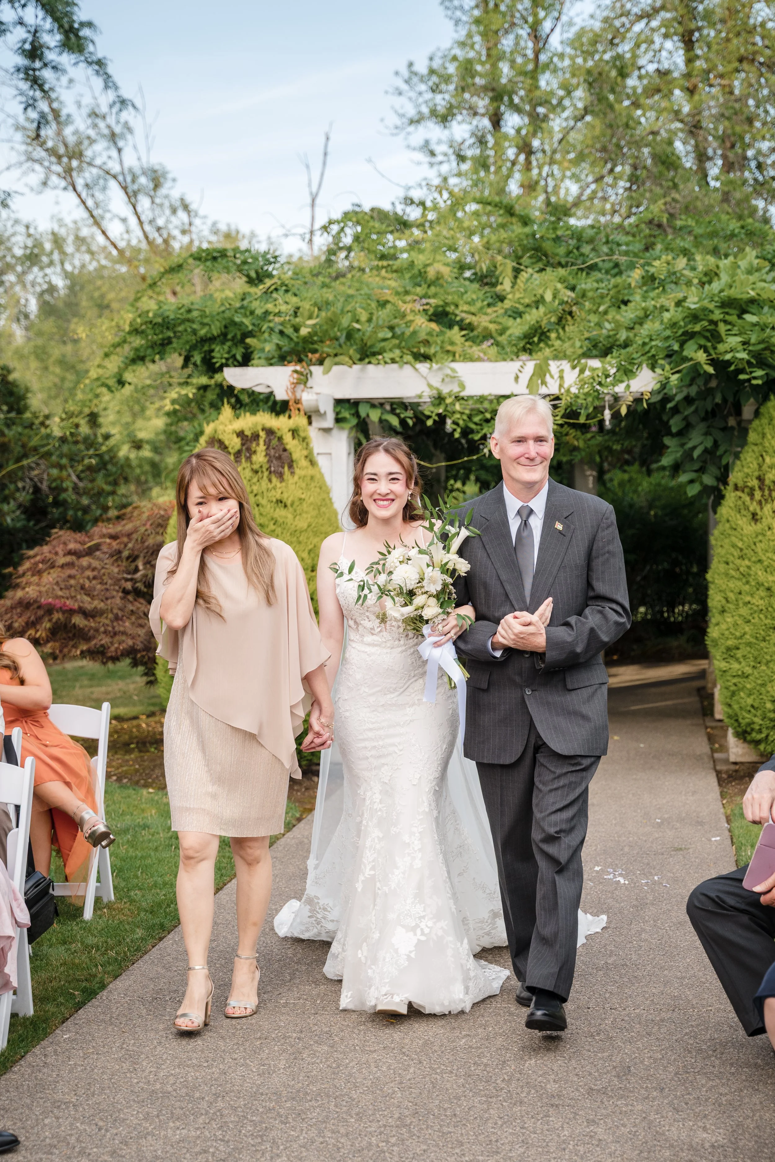 A bride in a white lace wedding dress holding a bouquet and walking arm-in-arm with an older man in a gray suit, while a woman laughs and covers her mouth on the same walk outdoors during daylight in a garden.