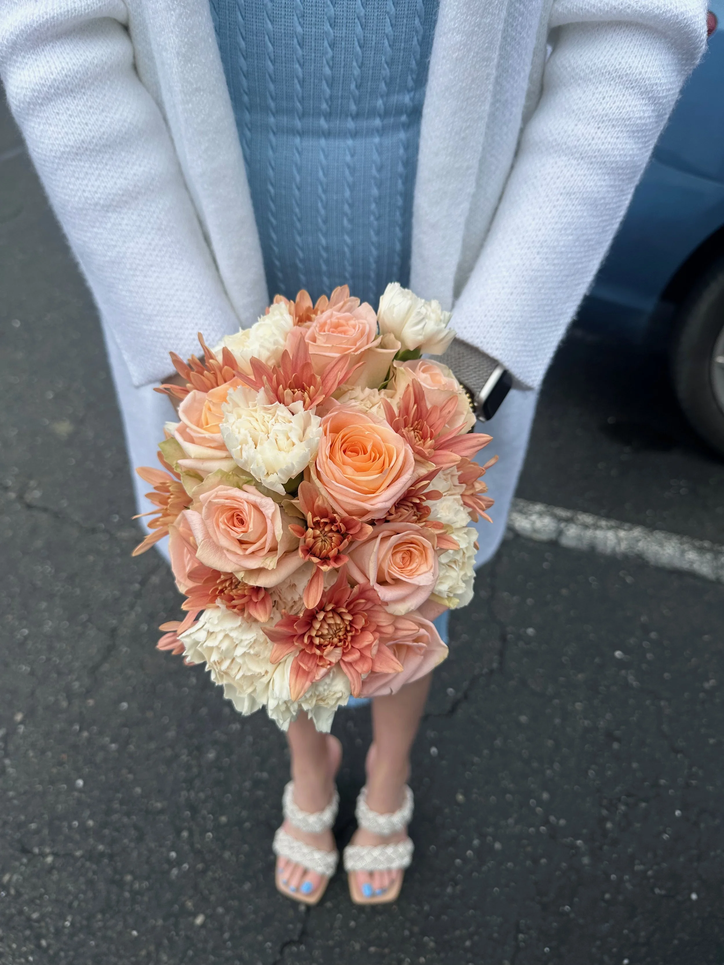 A person in a light sweater and sandals holds a bouquet of peach and cream-colored flowers, standing on a parking lot.
