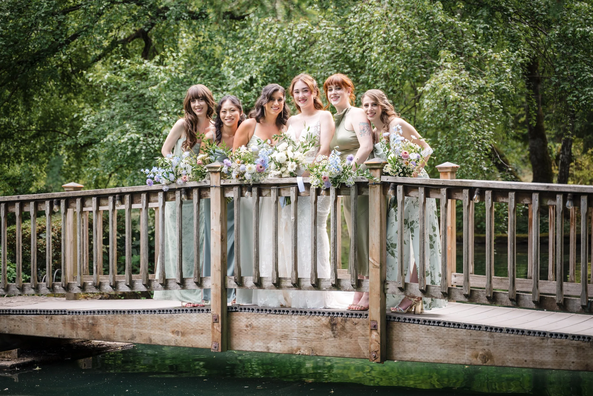 Group of women on a wooden bridge with flowers, surrounded by green trees.