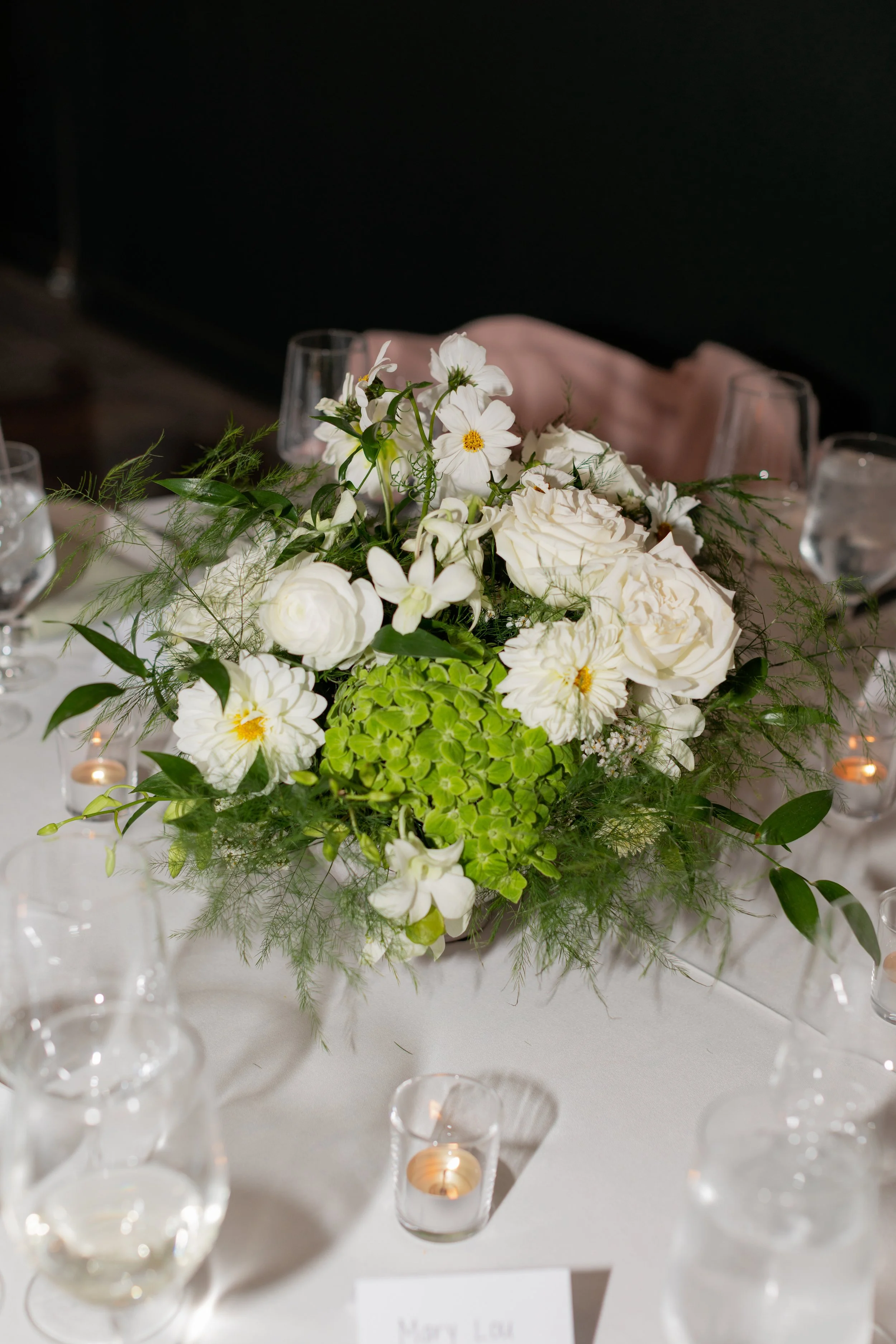 Elegant floral centerpiece with white roses, hydrangeas, and other white flowers on a table with candles in small glass holders.