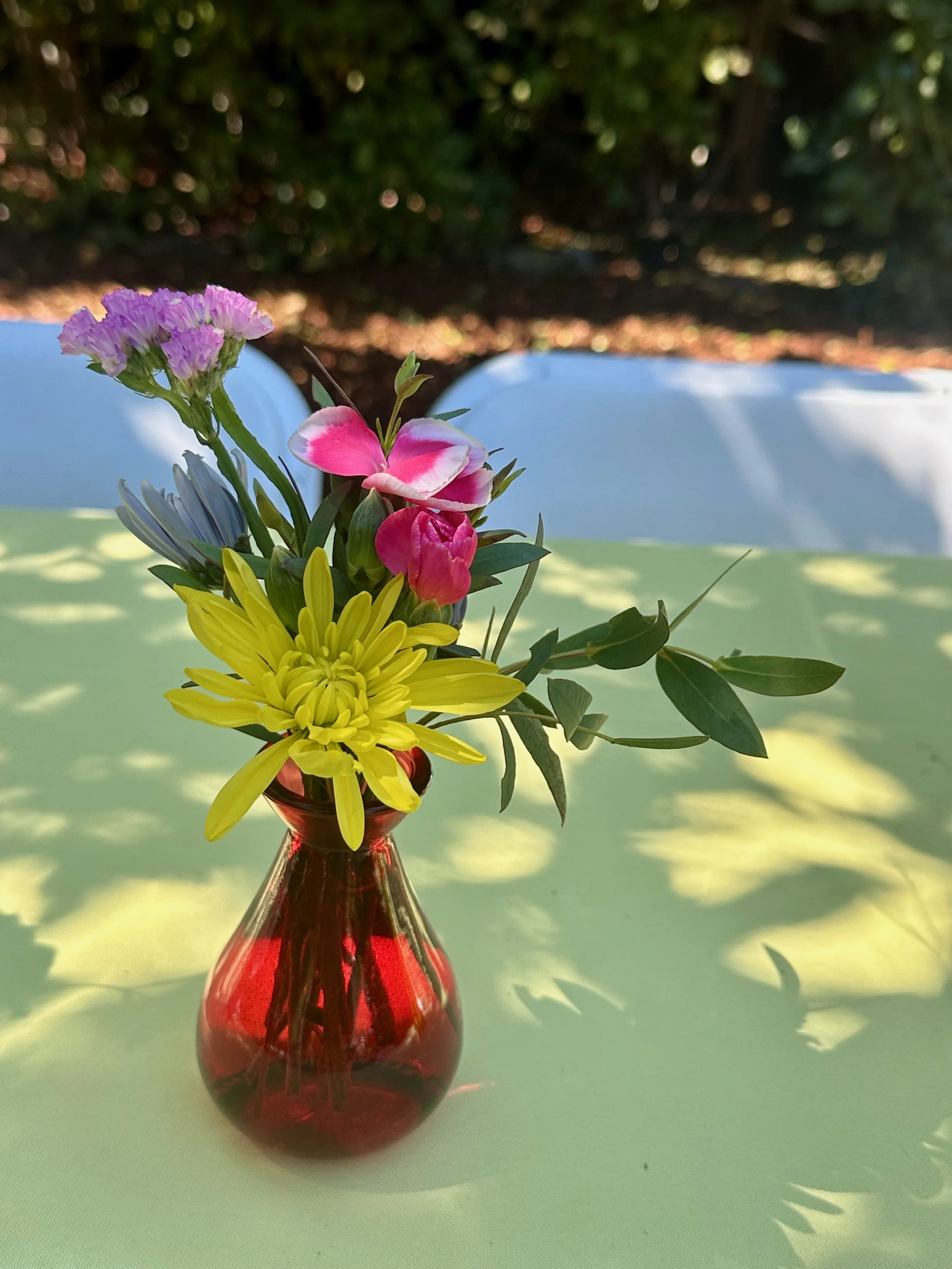 A small bouquet of multicolored flowers in a red glass vase on a light green tablecloth, with shadows and a blurred outdoor background.