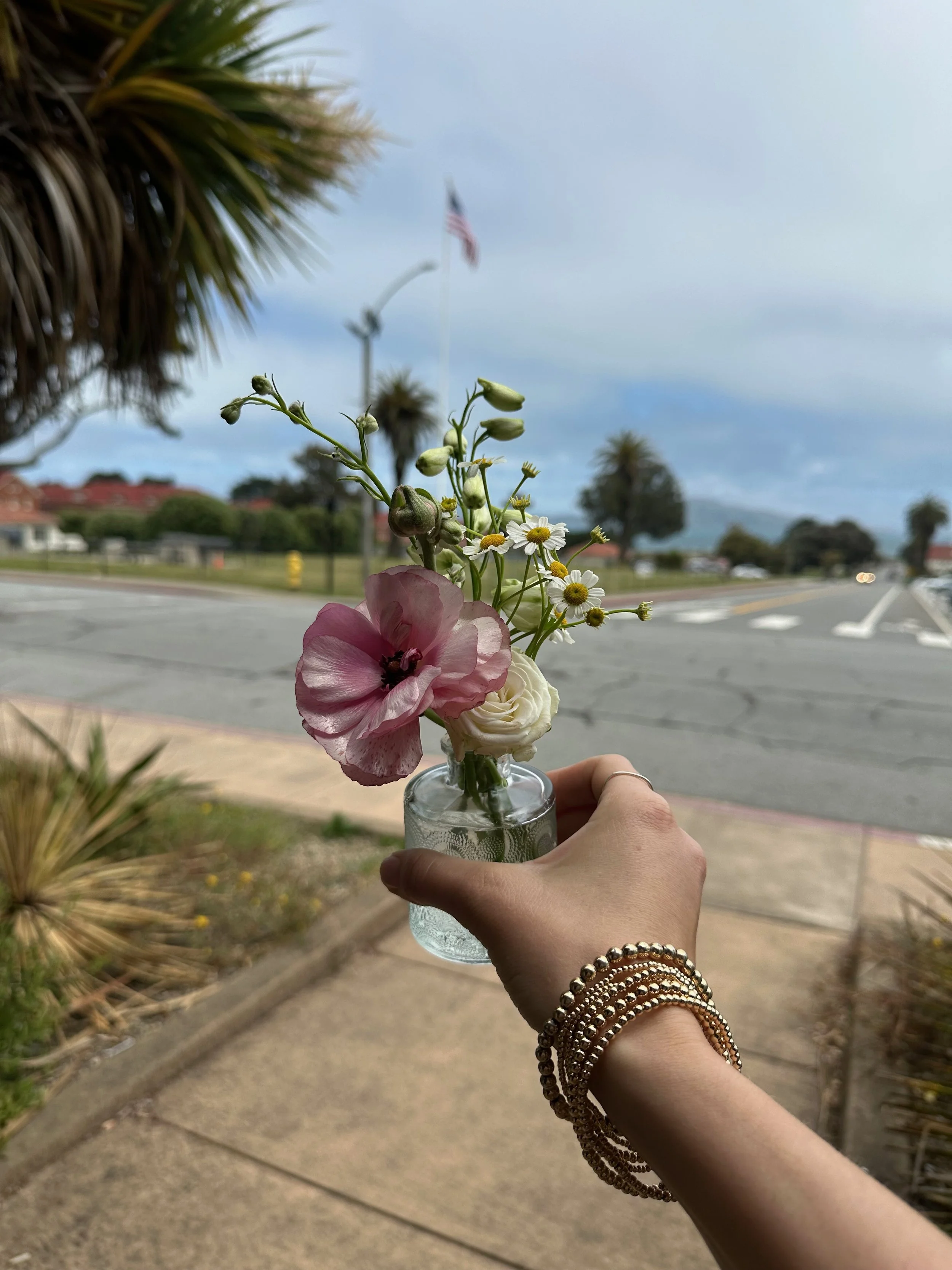 A hand holding a small glass jar with a bouquet of flowers, including pink, white, and small daisy-like flowers, with a street and cloudy sky in the background.