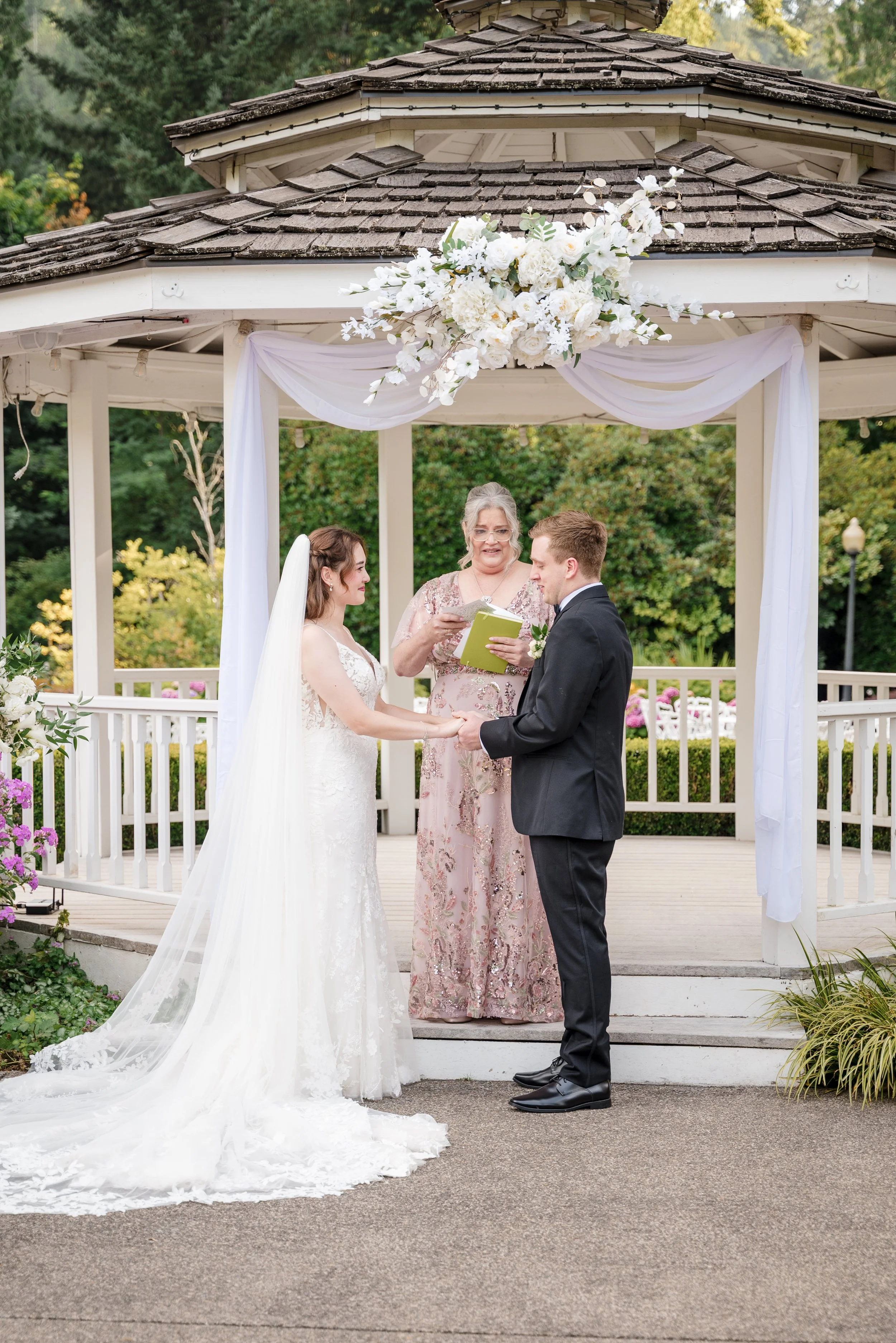 A bride and groom exchanging vows during their wedding ceremony beneath a decorated gazebo in a garden. The bride wears a white lace wedding gown and veil; the groom wears a black suit. An officiant, an older woman in a pastel pink dress, officiates 