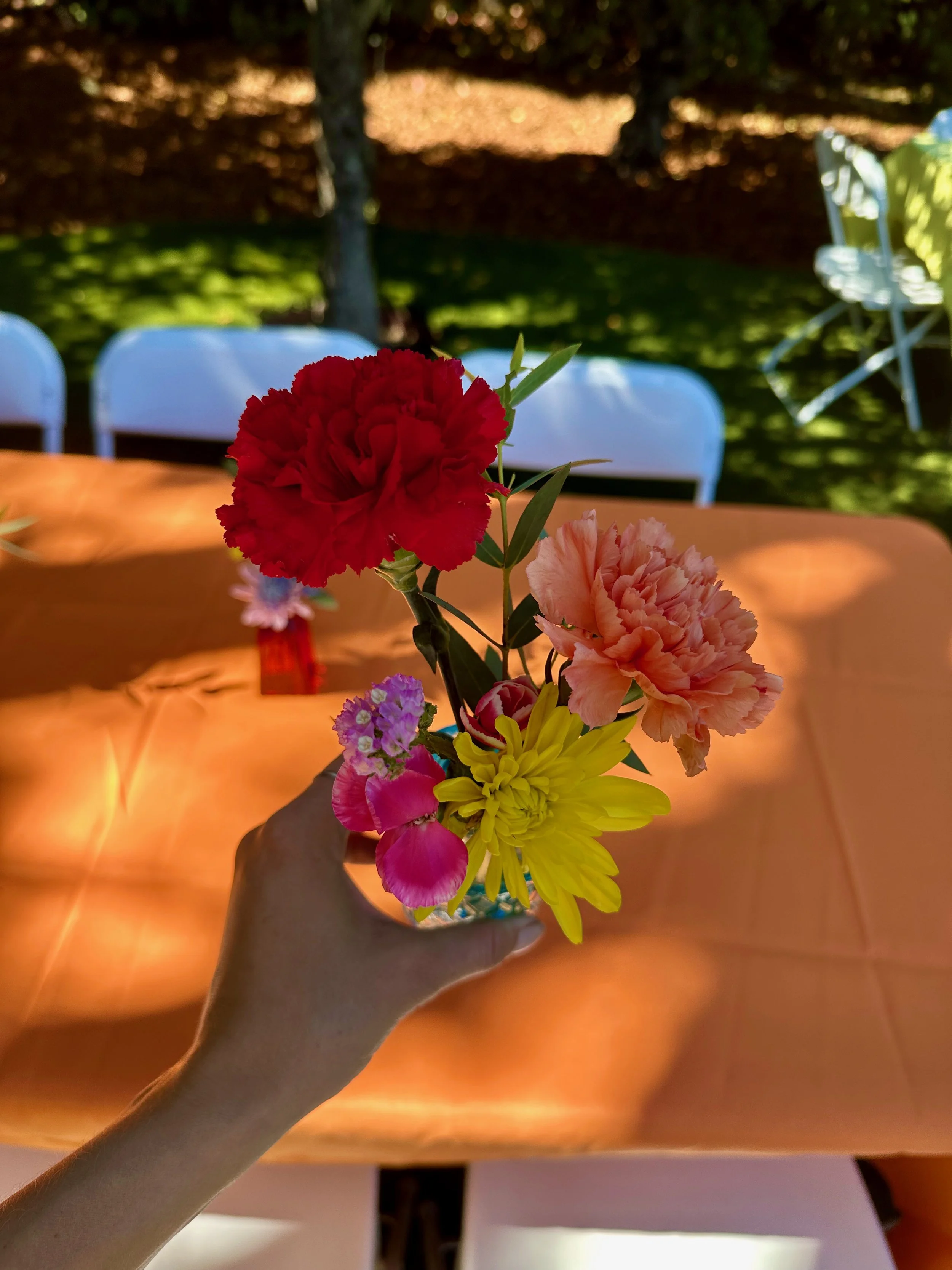 A hand holding a small bouquet of colorful flowers, including red, pink, yellow, and purple, with a blurred outdoor scene in the background.