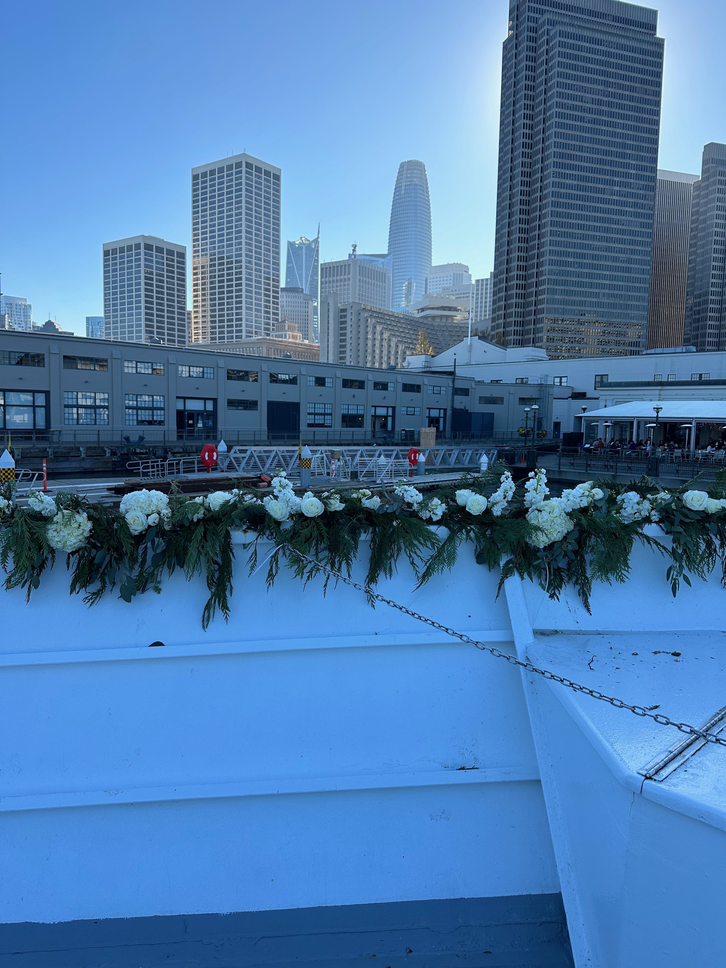 Cityscape of San Francisco skyline with tall skyscrapers, a waterfront with white floral and greenery decoration, and a dock in the foreground on a clear day.
