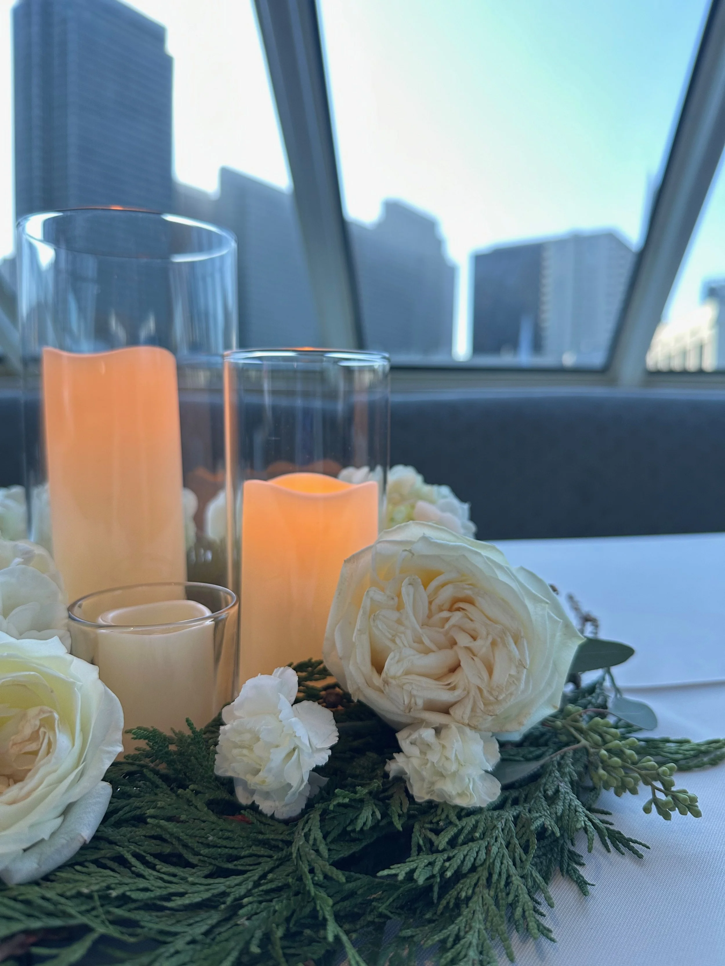 Elegant table centerpiece with white roses, greenery, and candles in glass holders, set against city skyscrapers visible through large windows.
