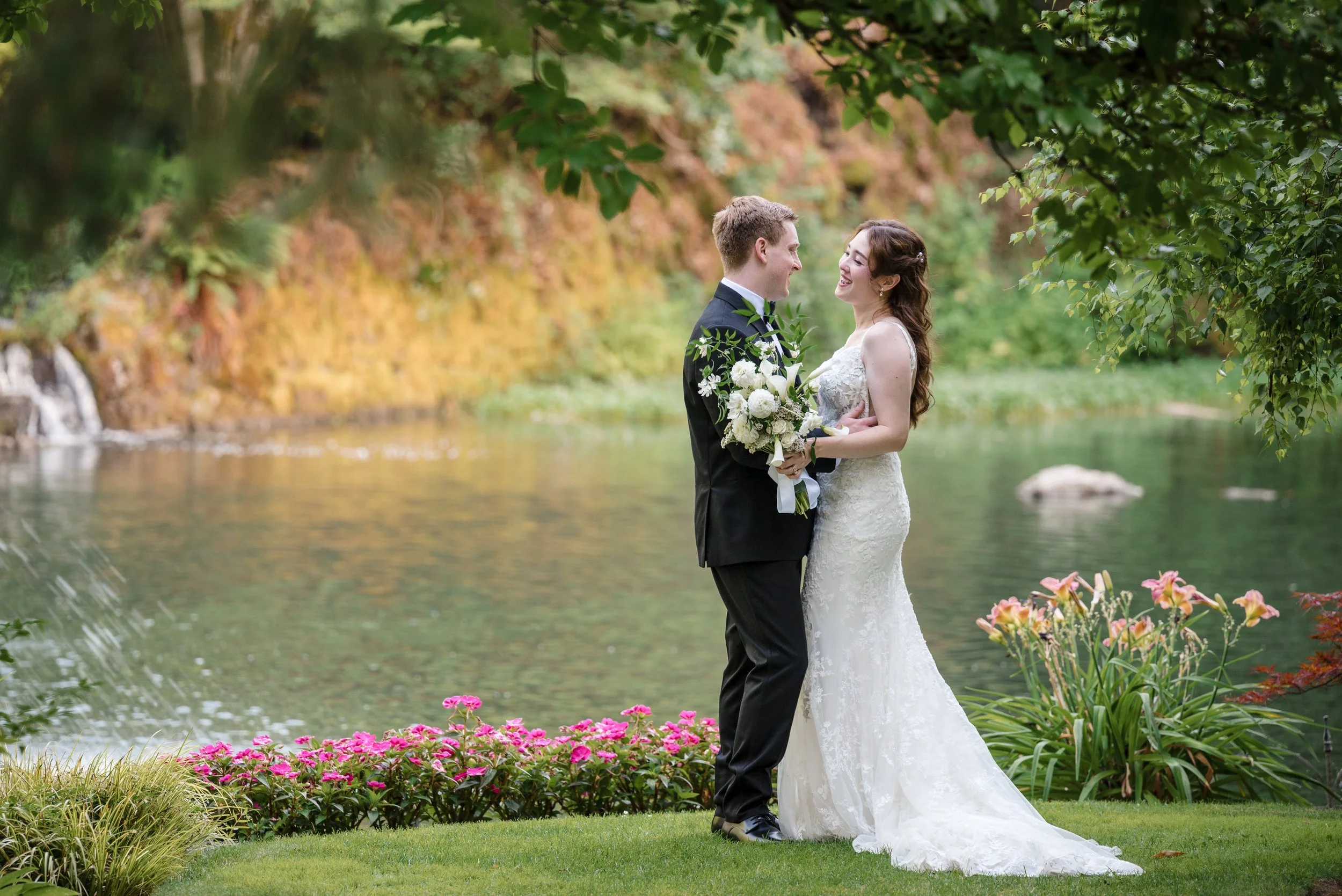 A bride and groom standing close together outdoors by a lake, smiling at each other. The bride is in a white wedding gown holding a bouquet, and the groom in a black tuxedo. Autumn foliage surrounds them.