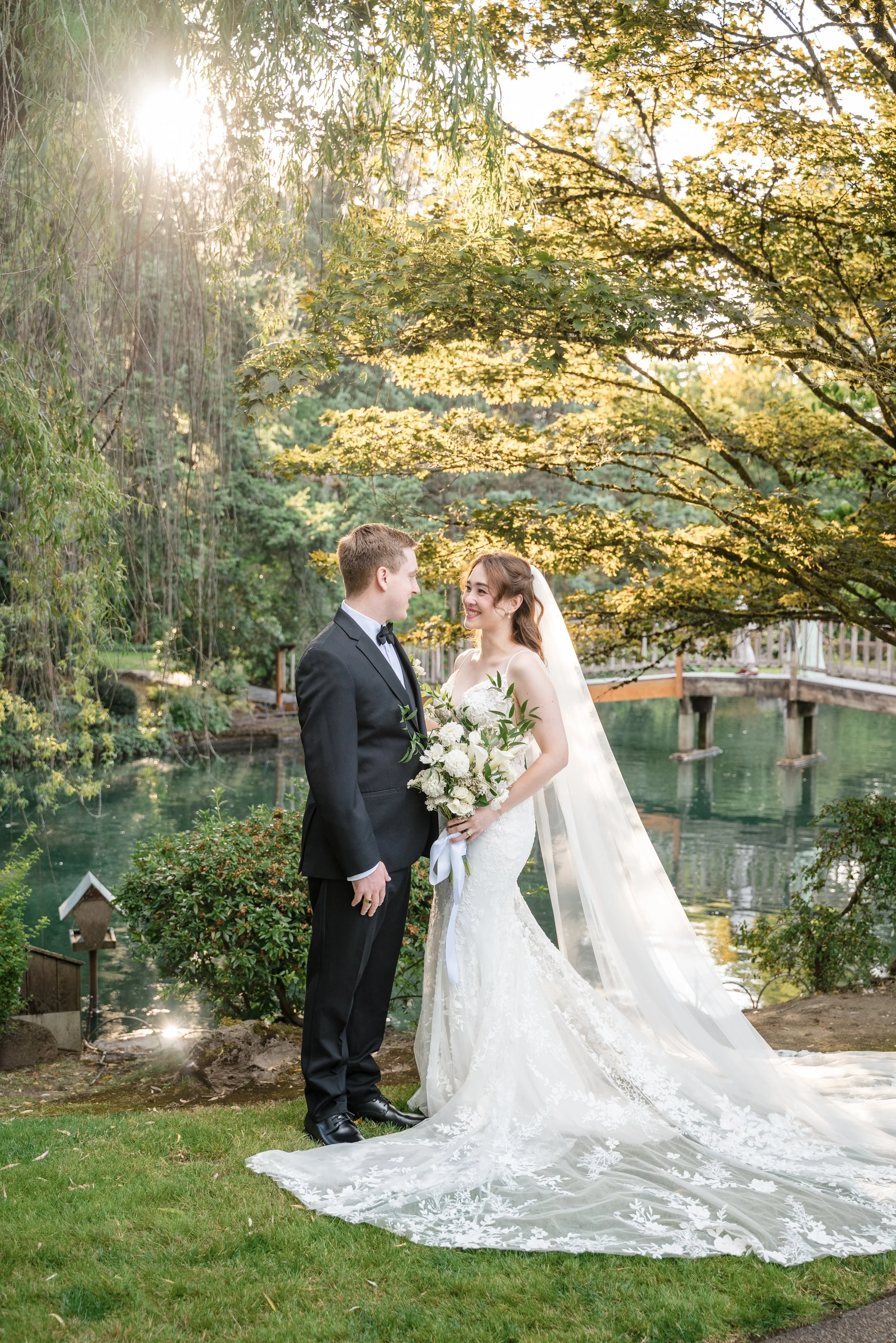 A bride and groom stand together outdoors by a pond, smiling at each other during sunset in a garden setting.