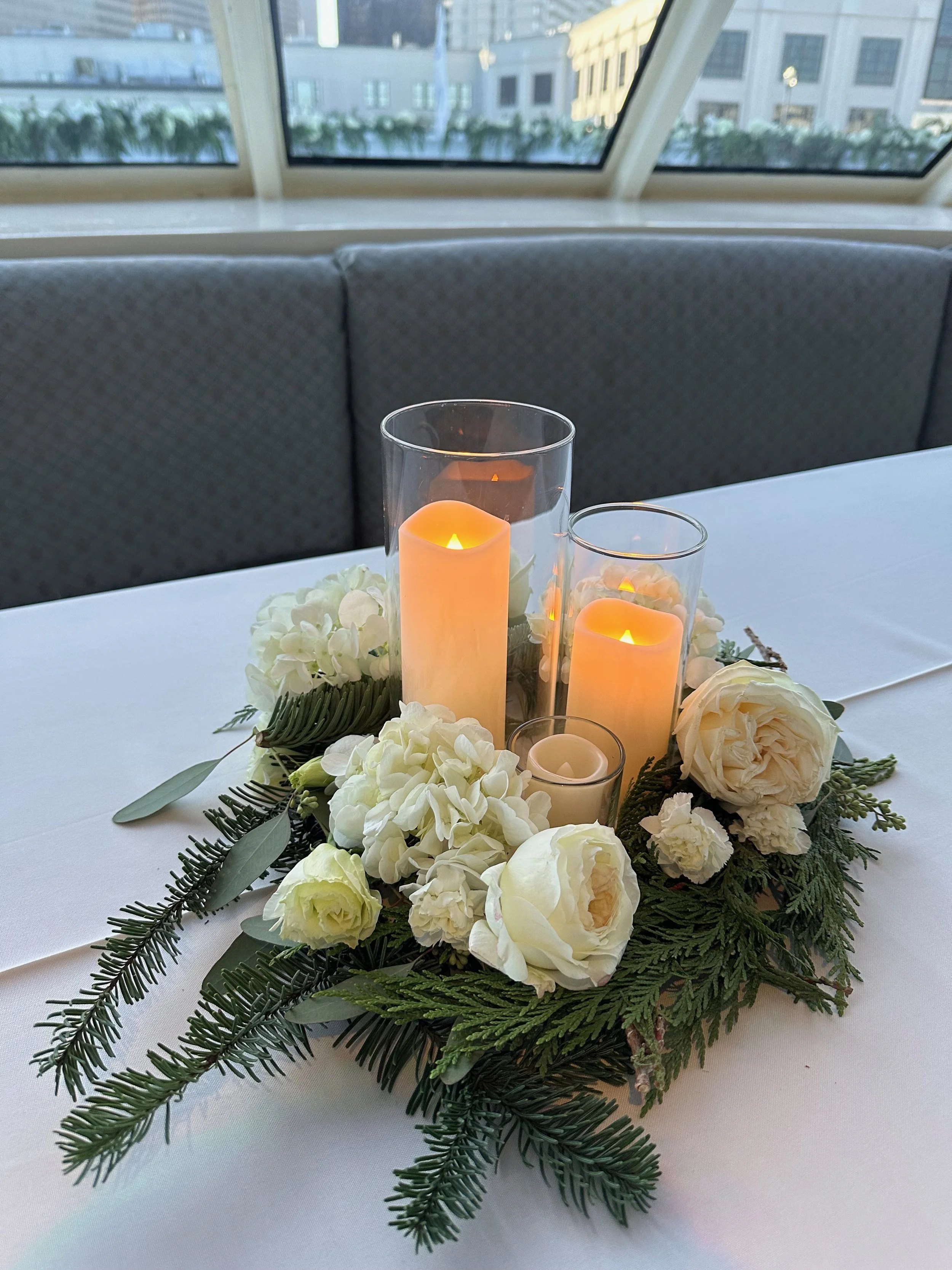 A floral centerpiece with white roses, hydrangeas, and greenery surrounding three lit candles in glass holders.