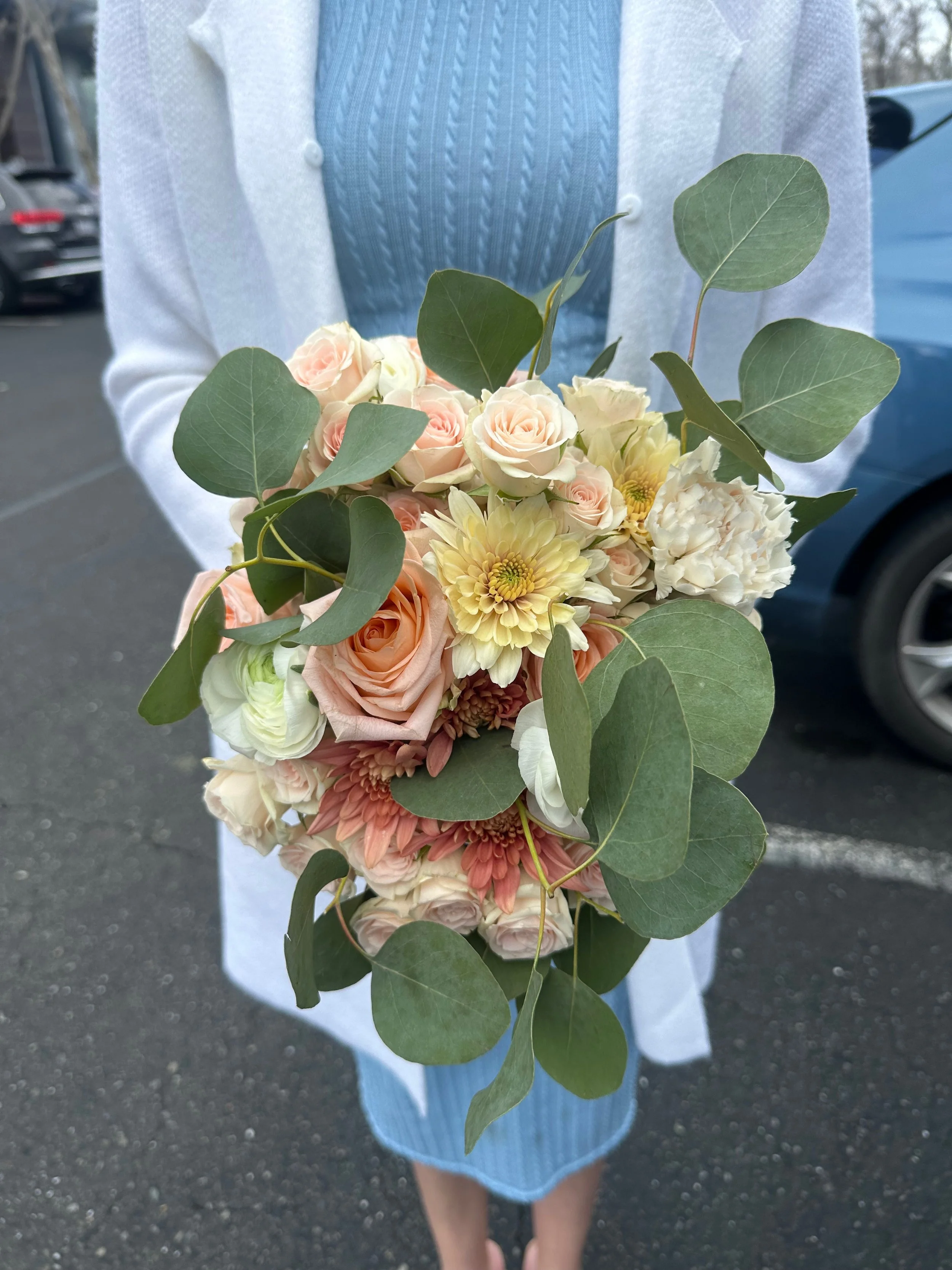 Person holding a bouquet of light pink roses, white and yellow chrysanthemums, and green eucalyptus leaves in a parking lot.