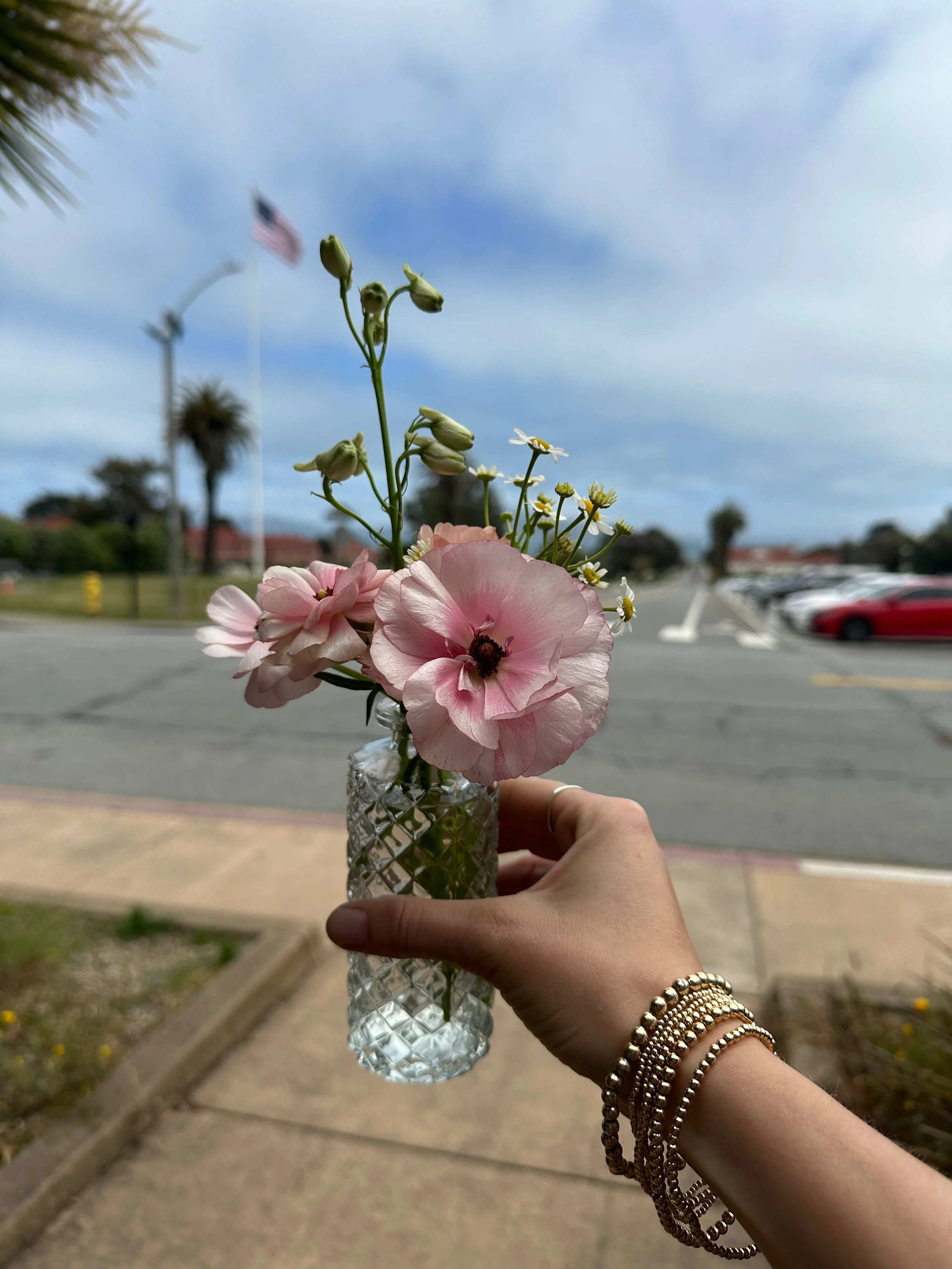 Person holding a glass vase with pink and white flowers on a sidewalk with parking lot and American flag in the background.