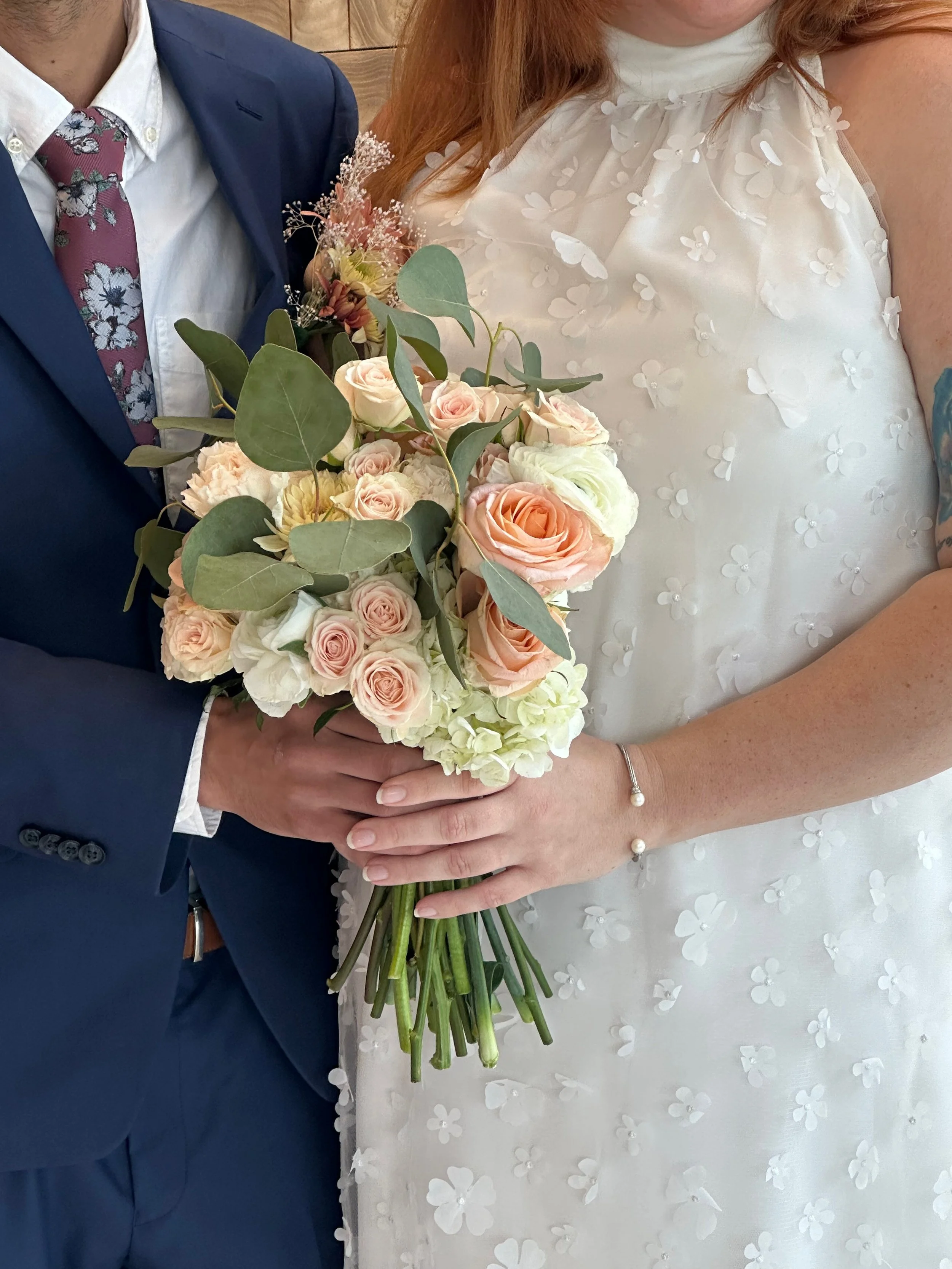 Close-up of a bride and groom holding a bouquet of pink and white roses, with the bride in a white dress with floral embellishments and the groom in a dark blue suit with a floral tie.