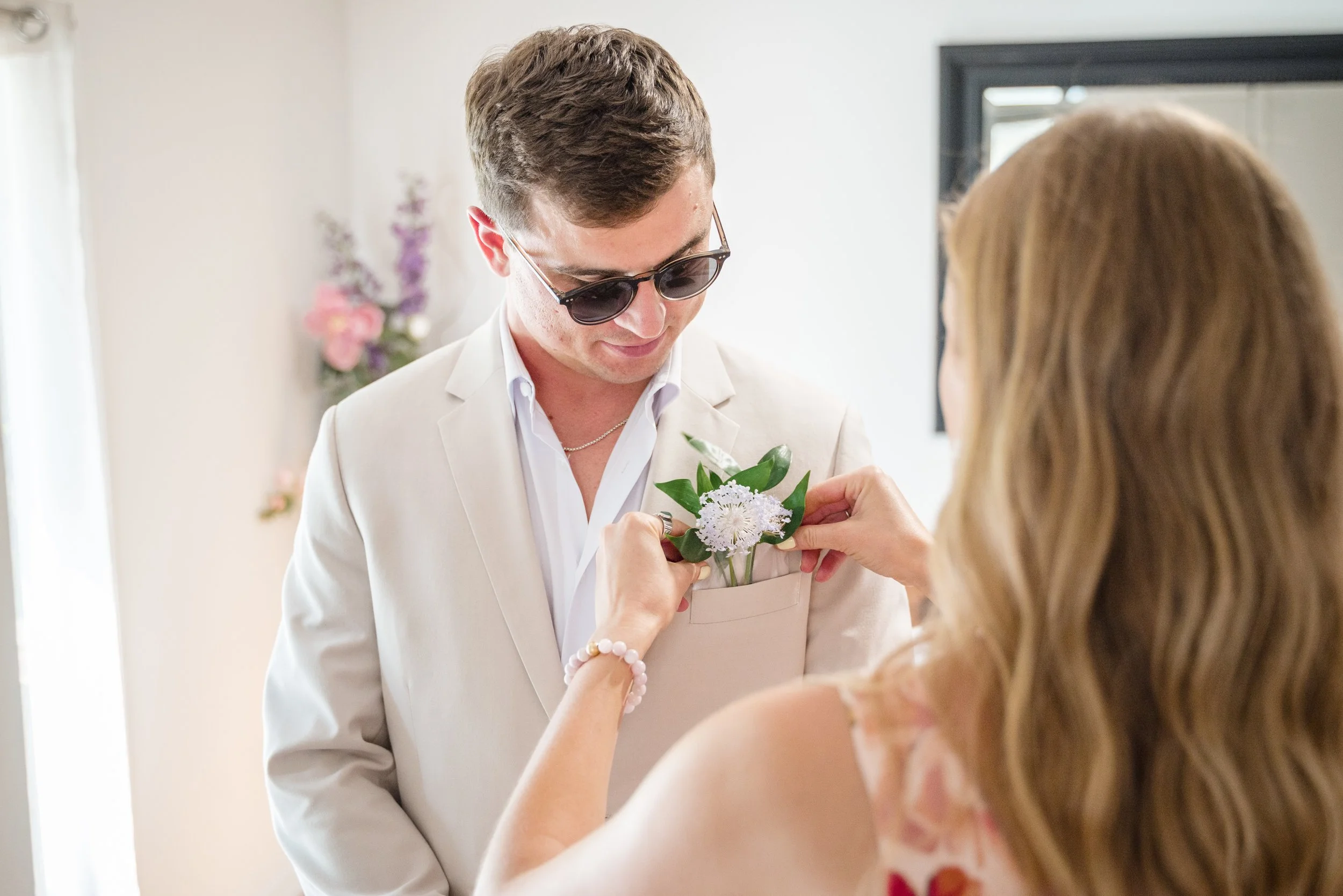 A man in a white suit and sunglasses receives a flower boutonniere from a woman during a wedding or special event.