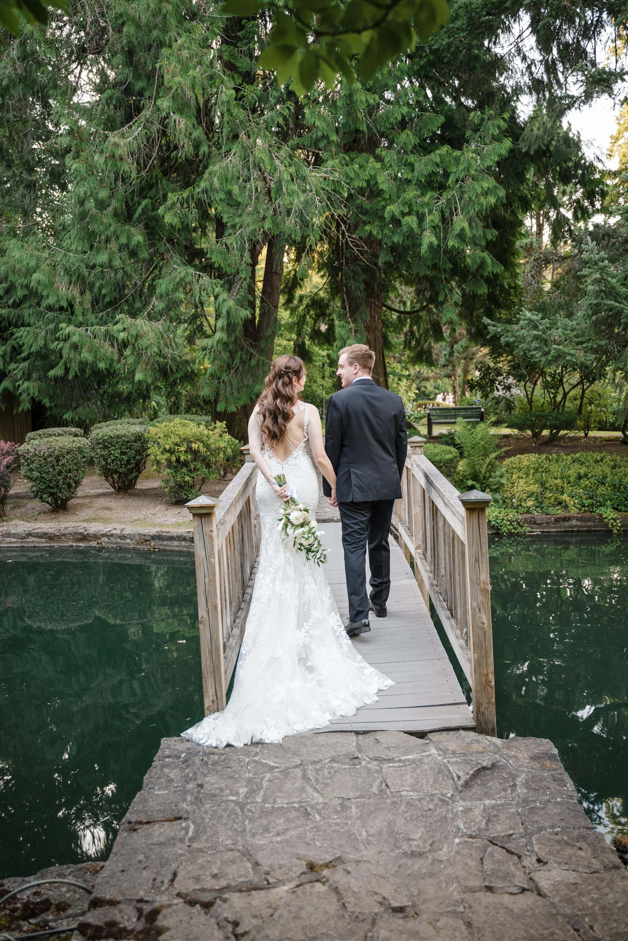 A bride and groom walk hand in hand across a small wooden bridge in a lush green park, surrounded by trees and shrubs during daytime.