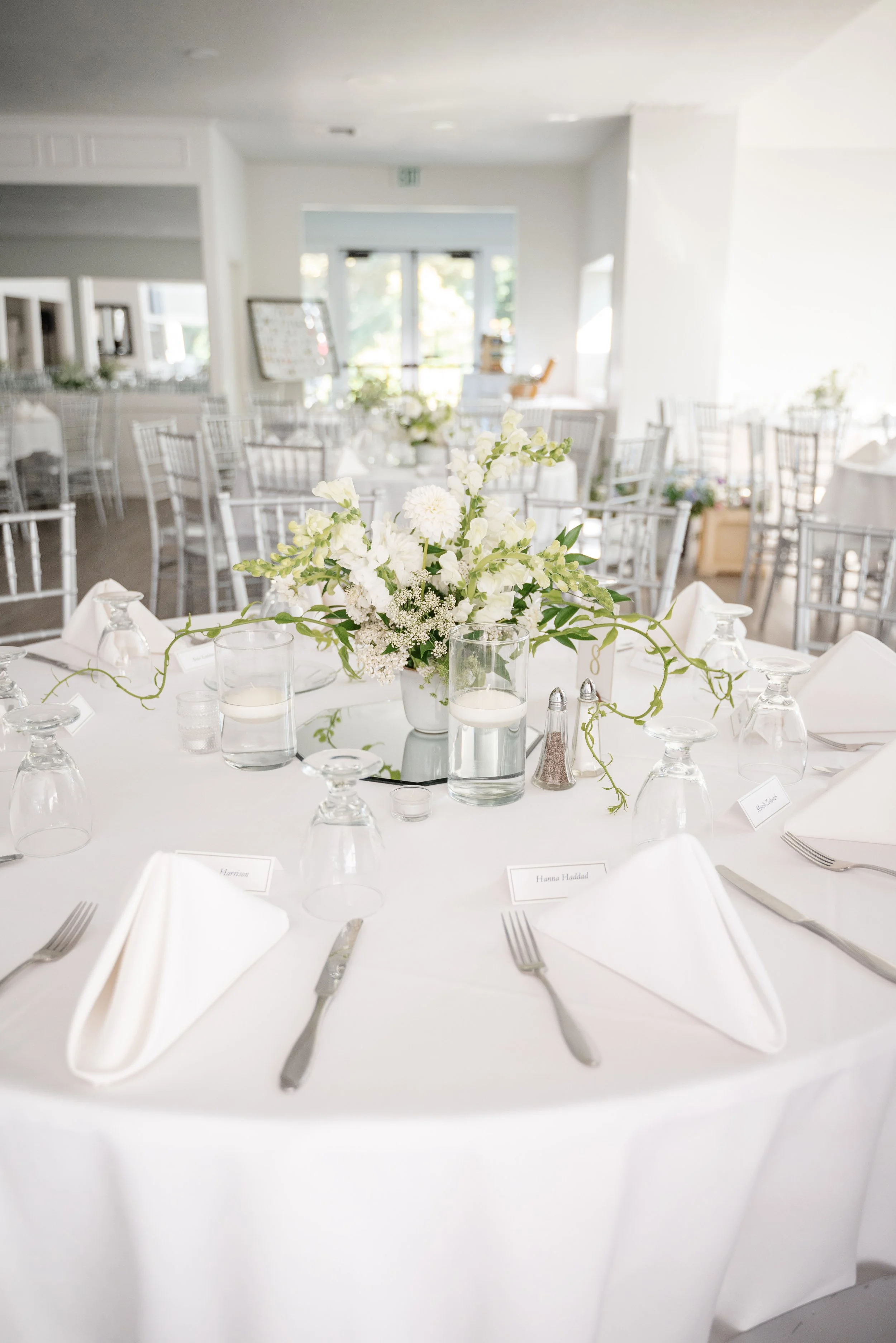 Wedding reception table with white tablecloth, floral centerpiece with white flowers and greenery, place settings with white napkins, silverware, upside-down glasses, water candles, and name cards in a bright, airy room.