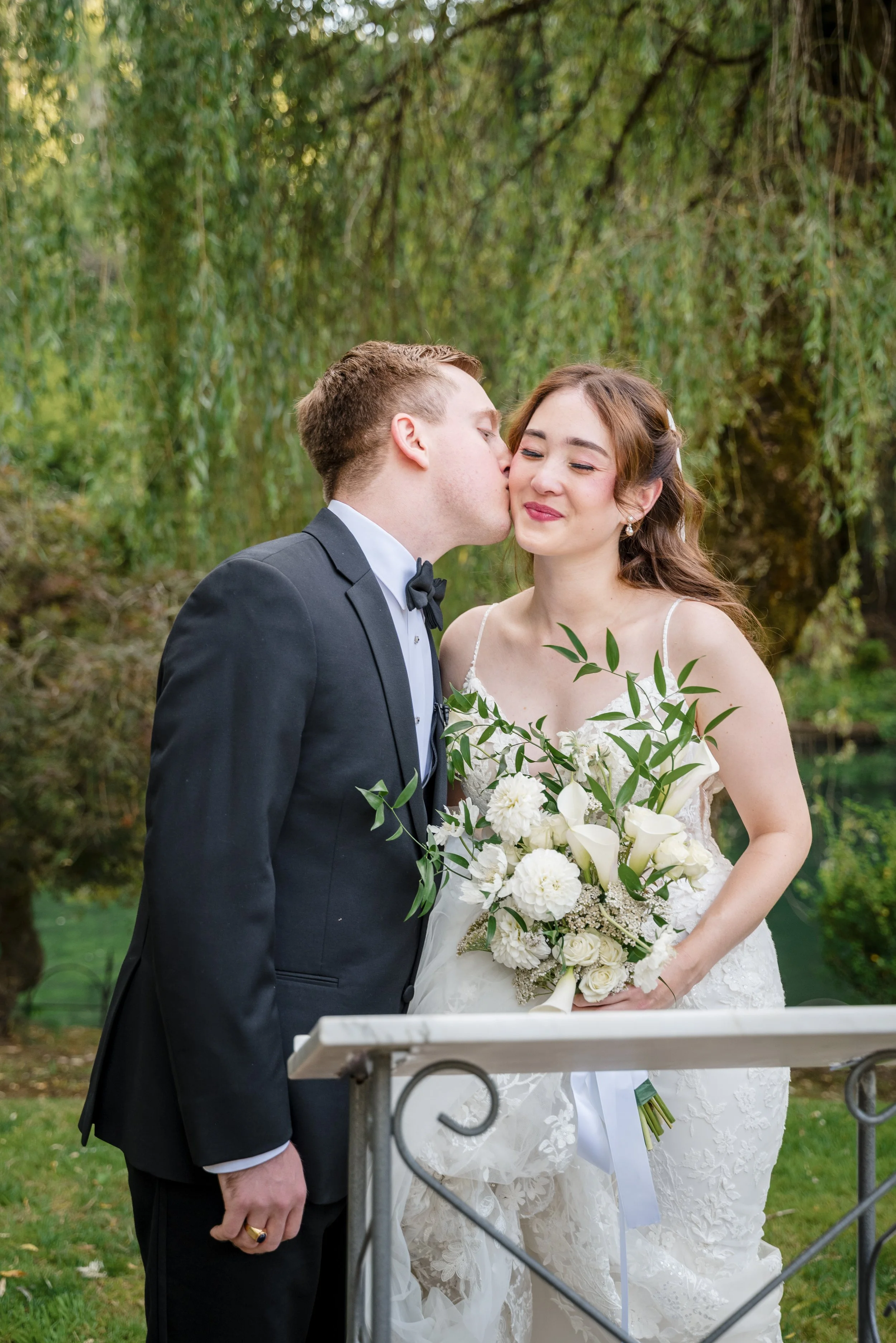 A groom in a black tuxedo and bow tie kisses a bride in a white wedding dress holding a bouquet of white flowers outdoors with green trees in the background.